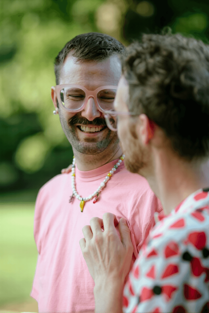 Two people smiling and chatting outdoors, one with pink glasses and a pink shirt with a beaded necklace, the other with glasses and a shirt with red and white patterns, in a lush green park.