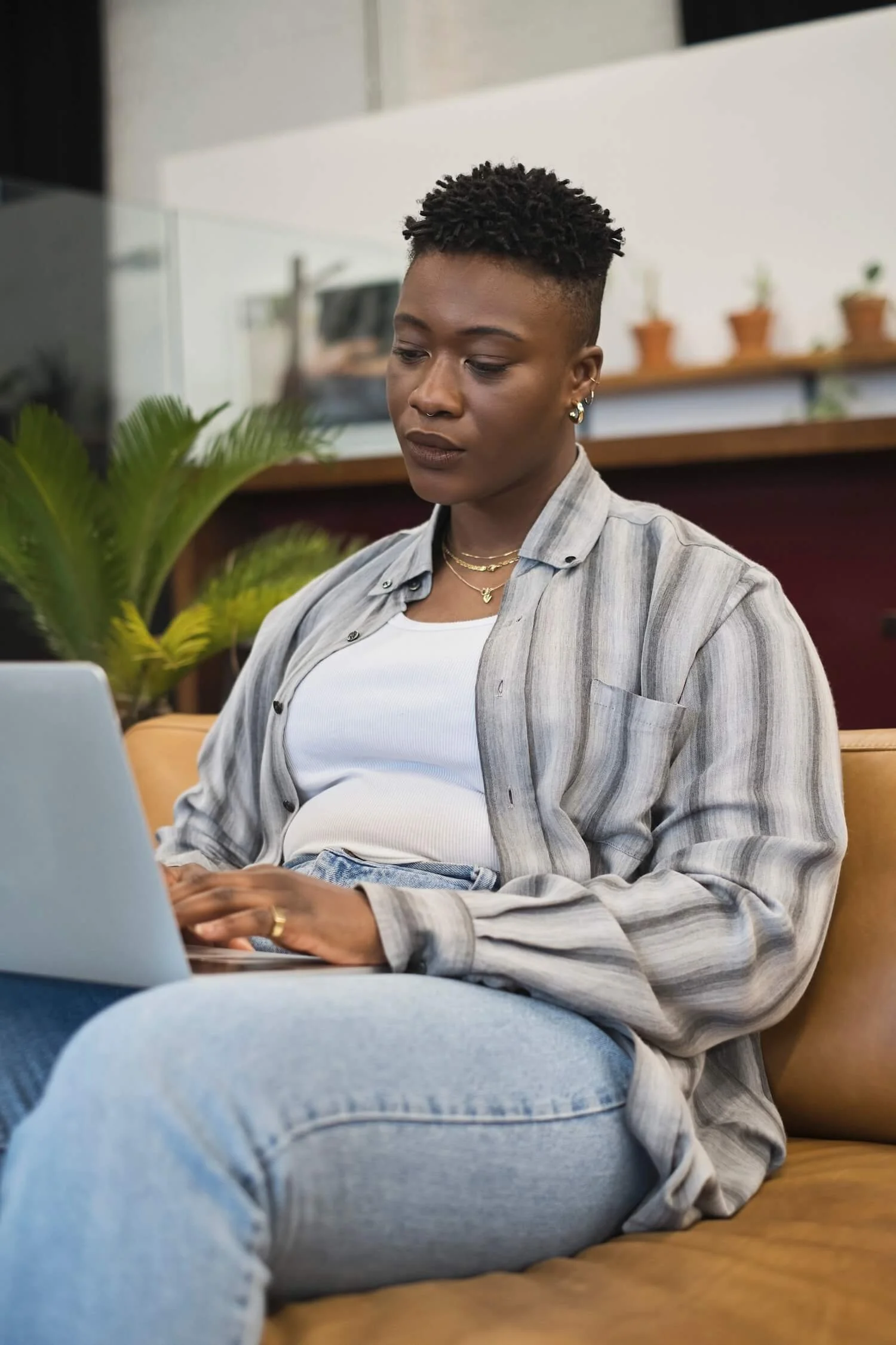 A non-binary person with short curly hair and multiple earrings sitting on a tan leather sofa, using a laptop in a modern indoor space with a plant and small potted plants in the background.