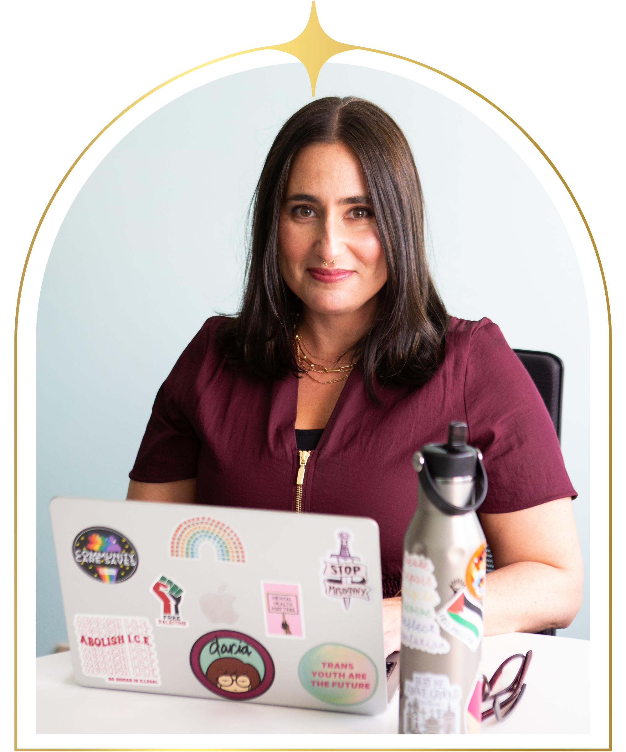 A woman with brown hair, wearing a burgundy top, sitting at a desk with a laptop decorated with various stickers, a water bottle, and a pair of glasses.