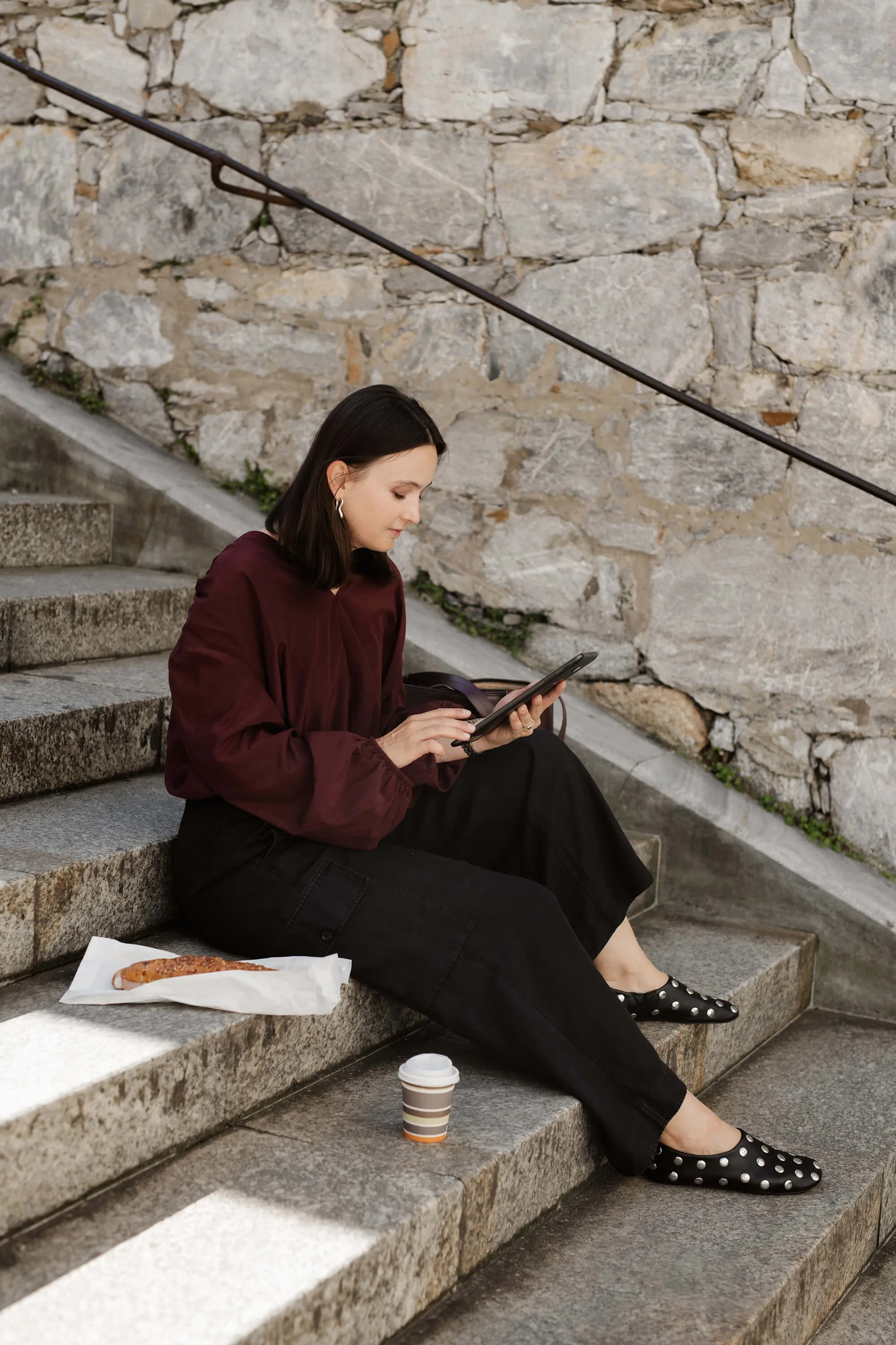 A woman sitting on stone stairs against a stone wall, using a tablet device, with a sandwich and coffee nearby.