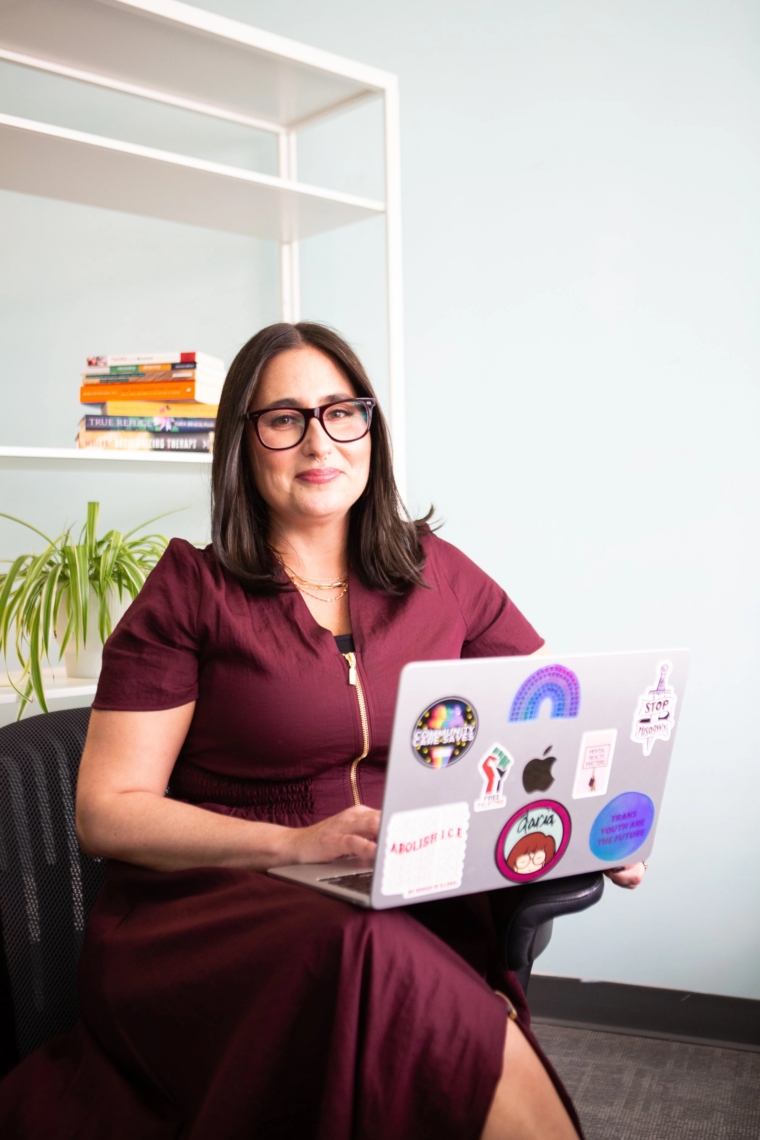 A woman with dark brown hair, wearing glasses and a maroon dress, sitting on a black office chair with a laptop covered in stickers, in an office with light walls, a bookshelf with visible books, and a green plant in the background.
