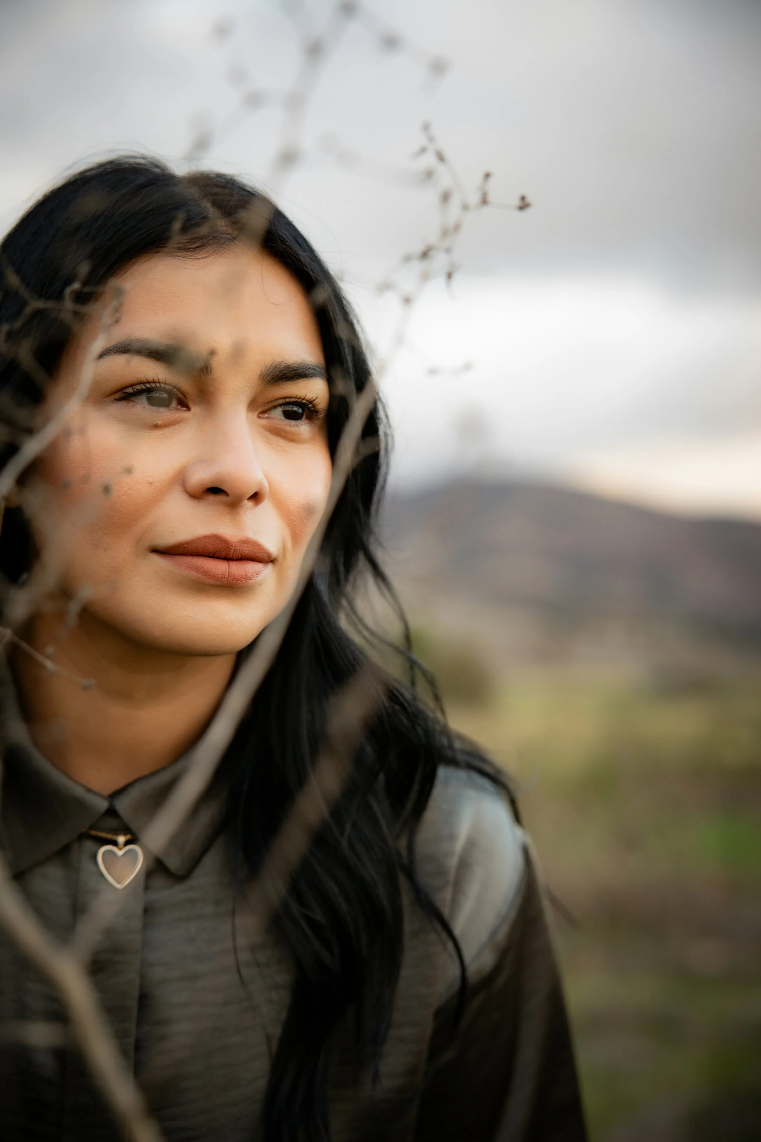 A woman with long black hair and light brown makeup looks off into the distance with a neutral expression outdoors, with branches partially obscuring the foreground and a blurred landscape in the background.
