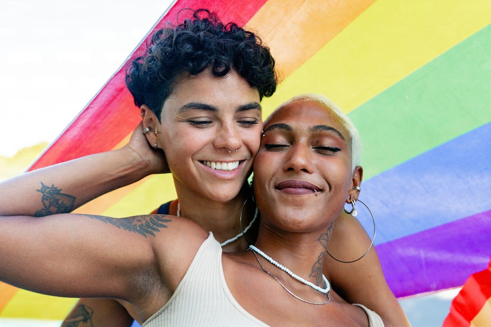 Two women with short hair and tattoos, smiling, embracing each other, in front of a rainbow pride flag.