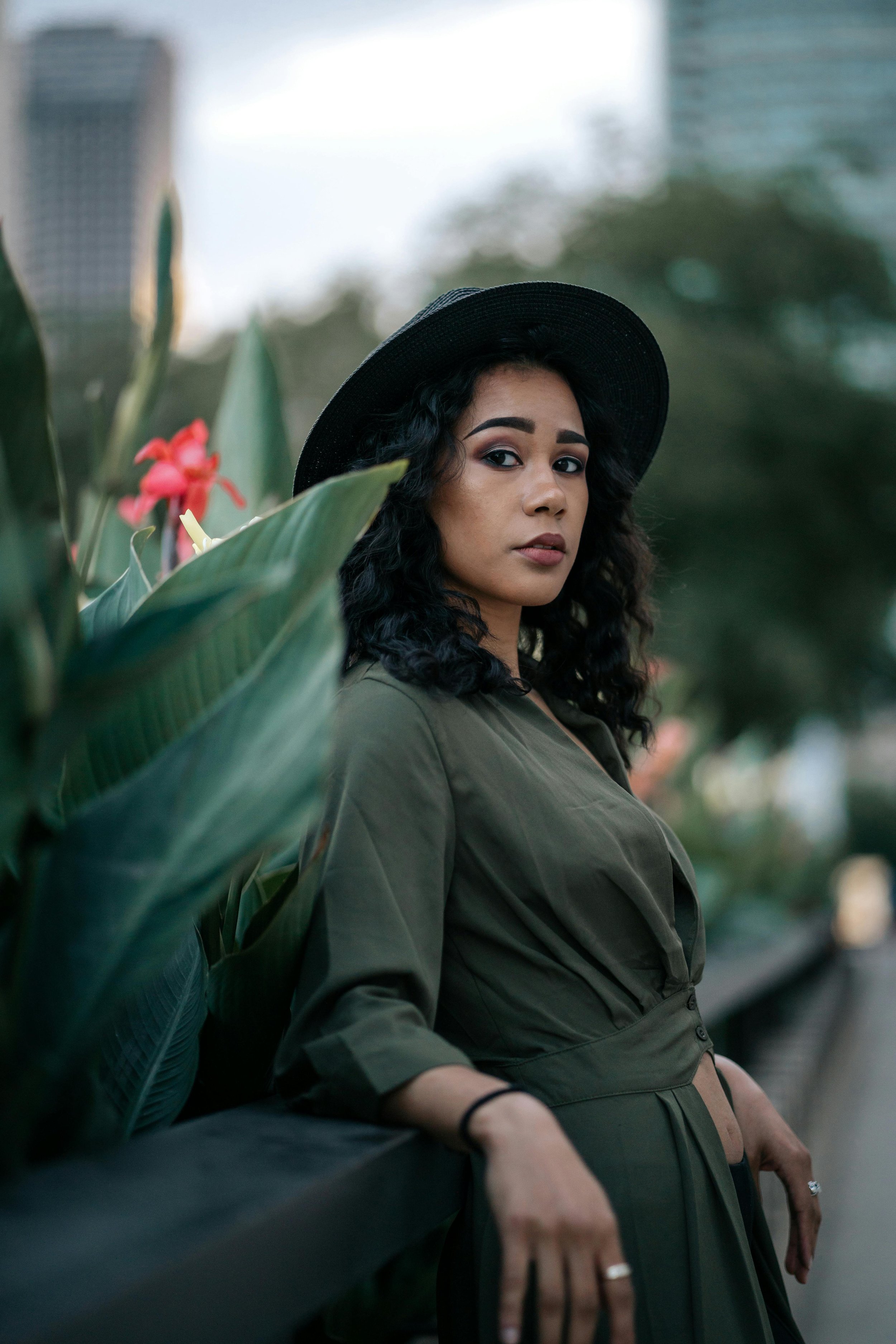 A woman with dark curly hair wearing a wide-brimmed black hat and an olive green dress, standing outdoors near large green plants, with a blurry city skyline in the background.