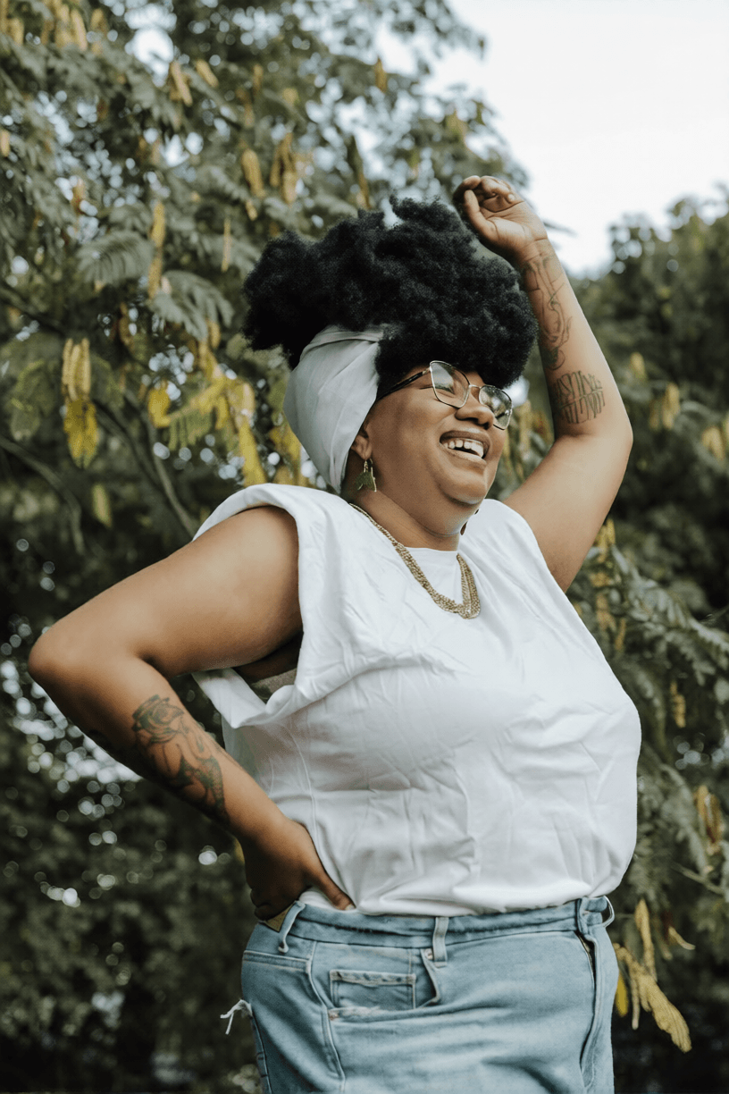A joyful woman with curly black hair pulled back with a white head wrap, wearing glasses, a white sleeveless top, and light blue jeans, standing outdoors with trees in the background.