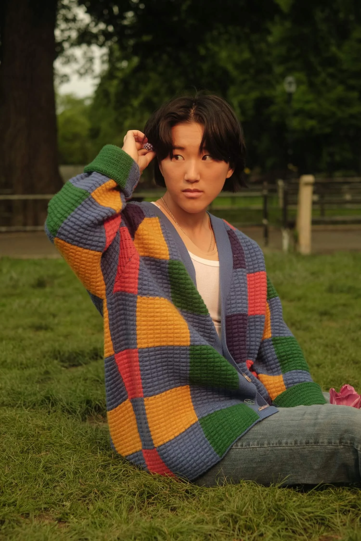 A non-binary trans man with short black hair sitting on grass in a park, wearing a colorful, checkered, textured sweater, a white undershirt, and gray jeans, looking to the side with a neutral expression.