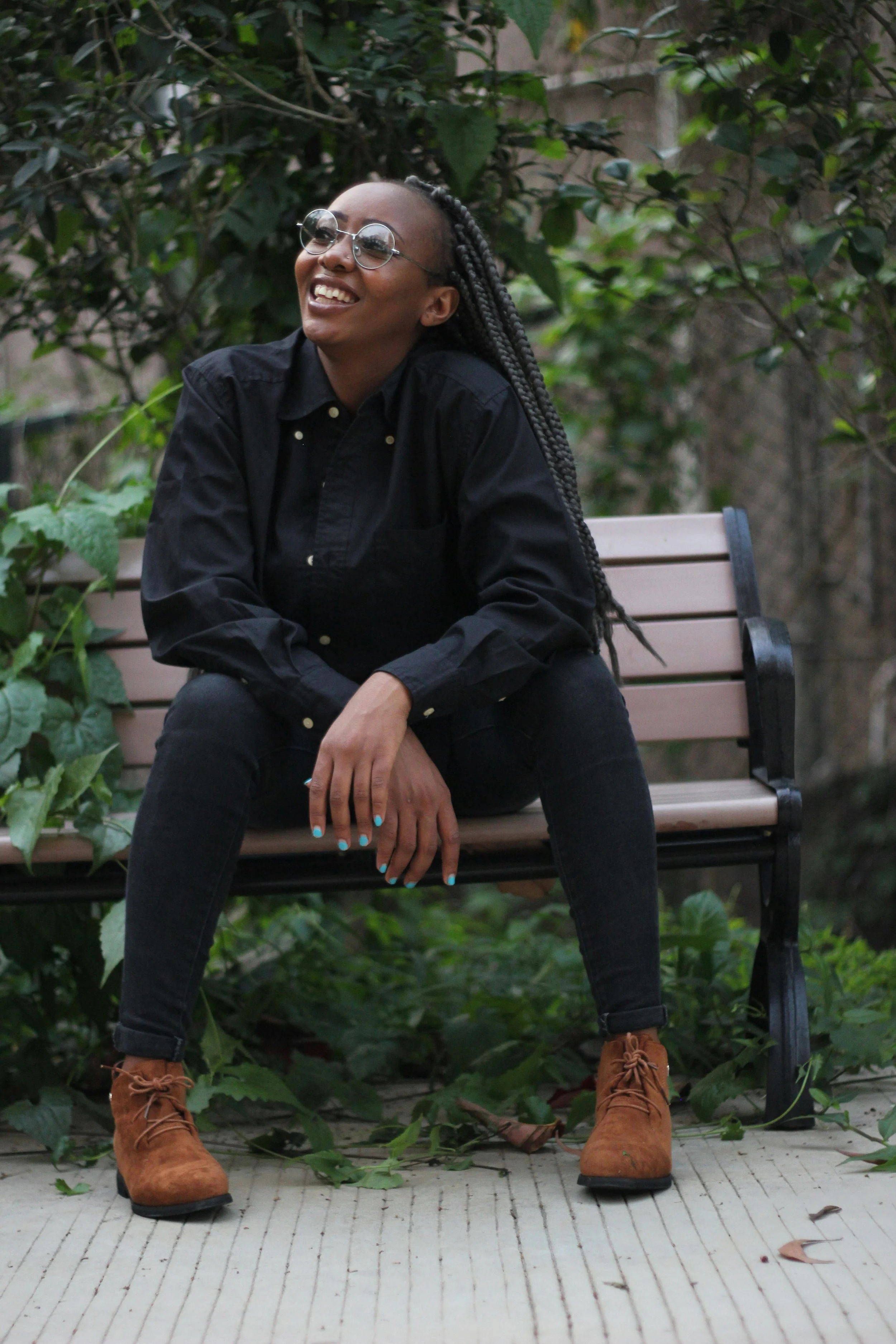A woman with long braided hair, wearing glasses, a black shirt, black pants, and brown boots, sitting on a park bench, smiling, with green foliage in the background.