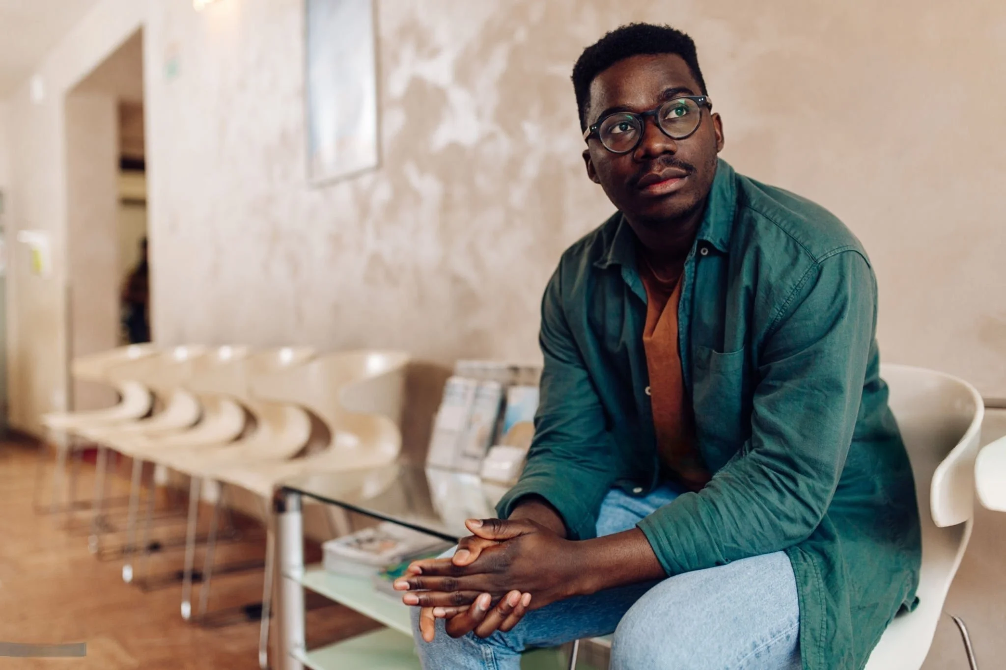 A young man with glasses and dark hair sits in a waiting area, looking off to the side with a thoughtful expression. He wears a green jacket over a brown shirt and light blue jeans. The background shows a row of empty chairs, a beige wall, and a glass table.