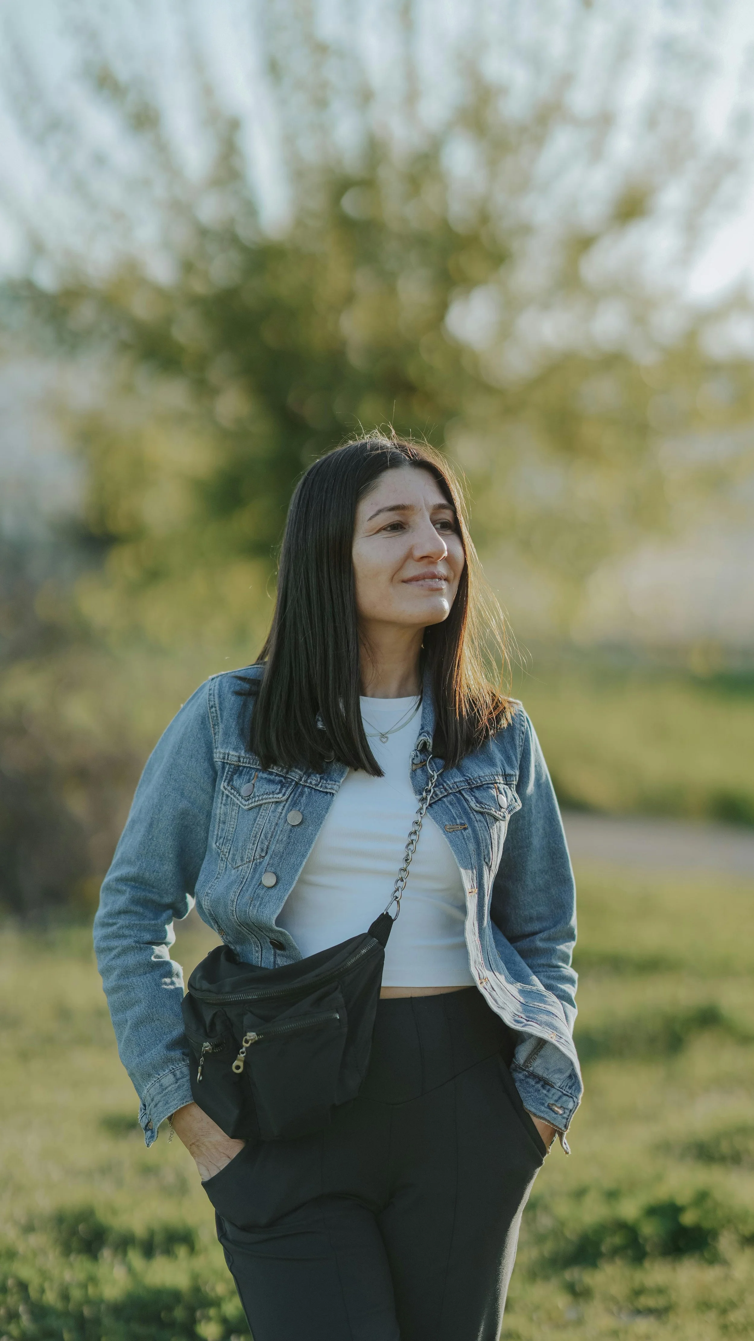 A woman walking outdoors with trees in the background, wearing a denim jacket, white t-shirt, black pants, and carrying a black crossbody bag.