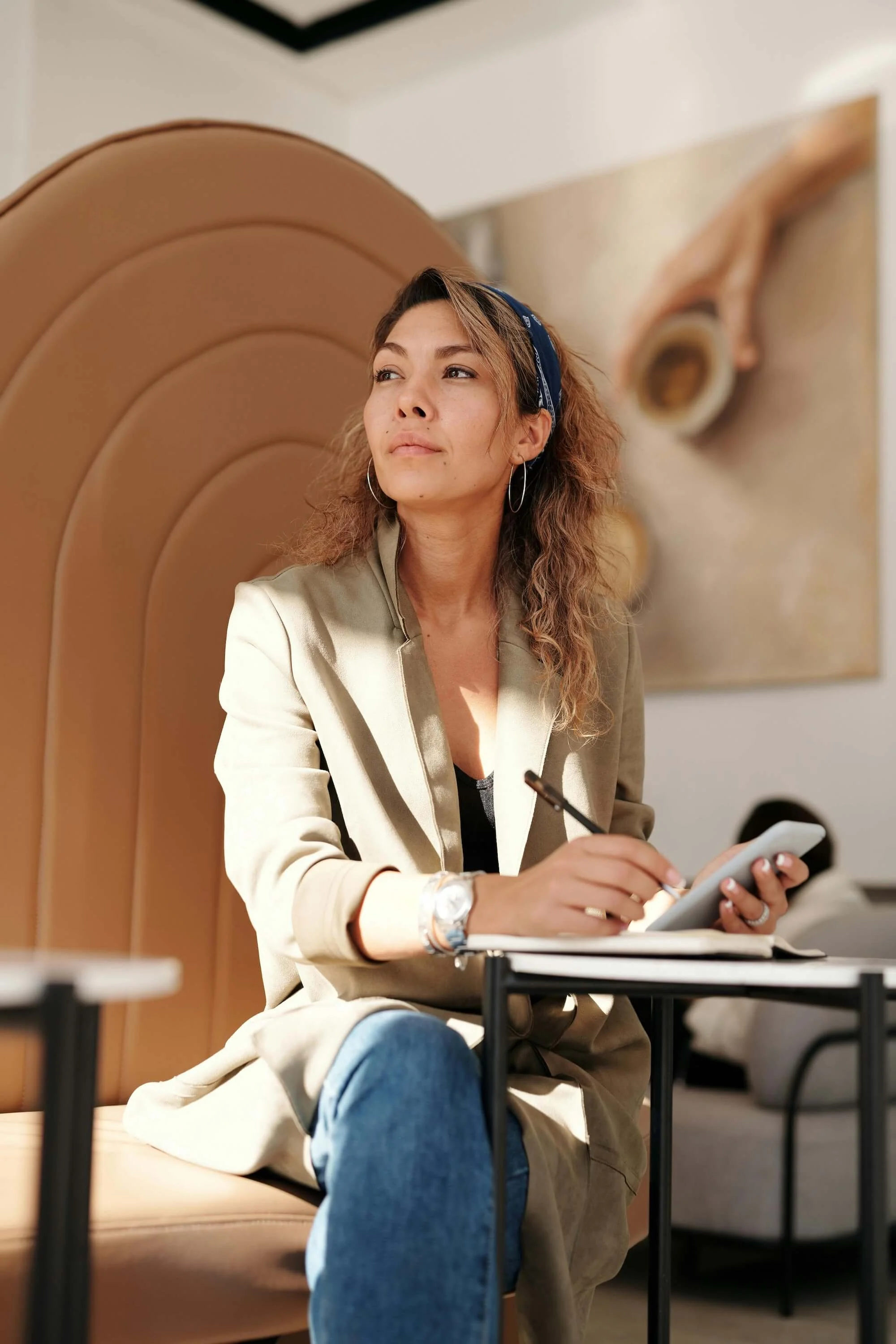 A woman with wavy hair wearing a beige blazer, headband, and hoop earrings, sitting on a beige chair with a high backrest. She is holding a pen and a notepad and looking to her left in a room with abstract wall art.