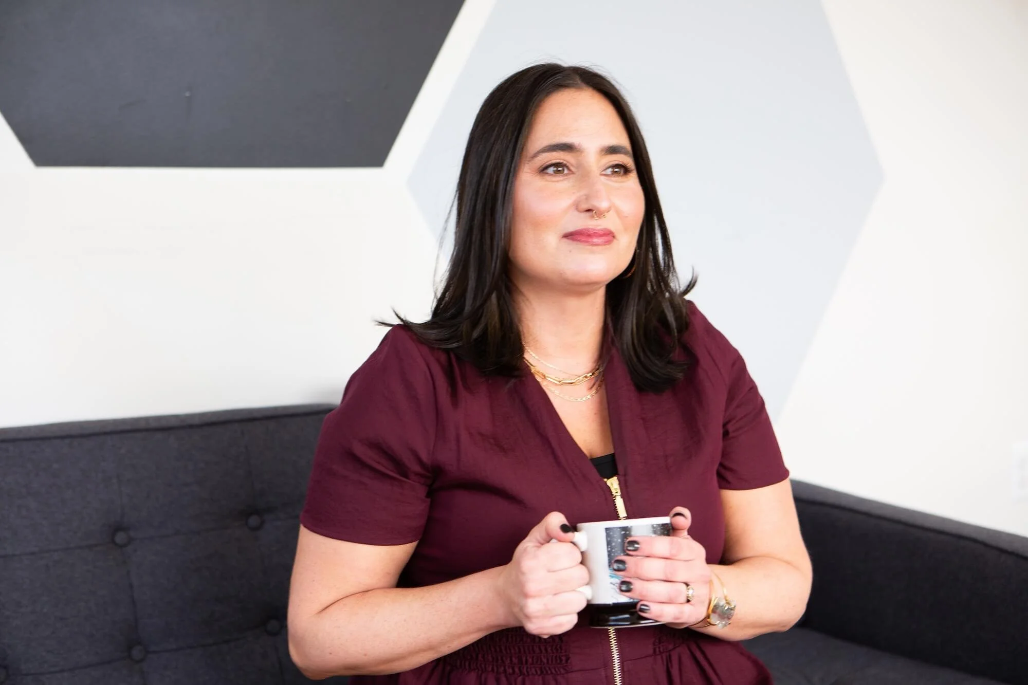 A woman with black hair, wearing a maroon top, holding a coffee mug, sitting on a dark grey couch in front of a geometric patterned wall.