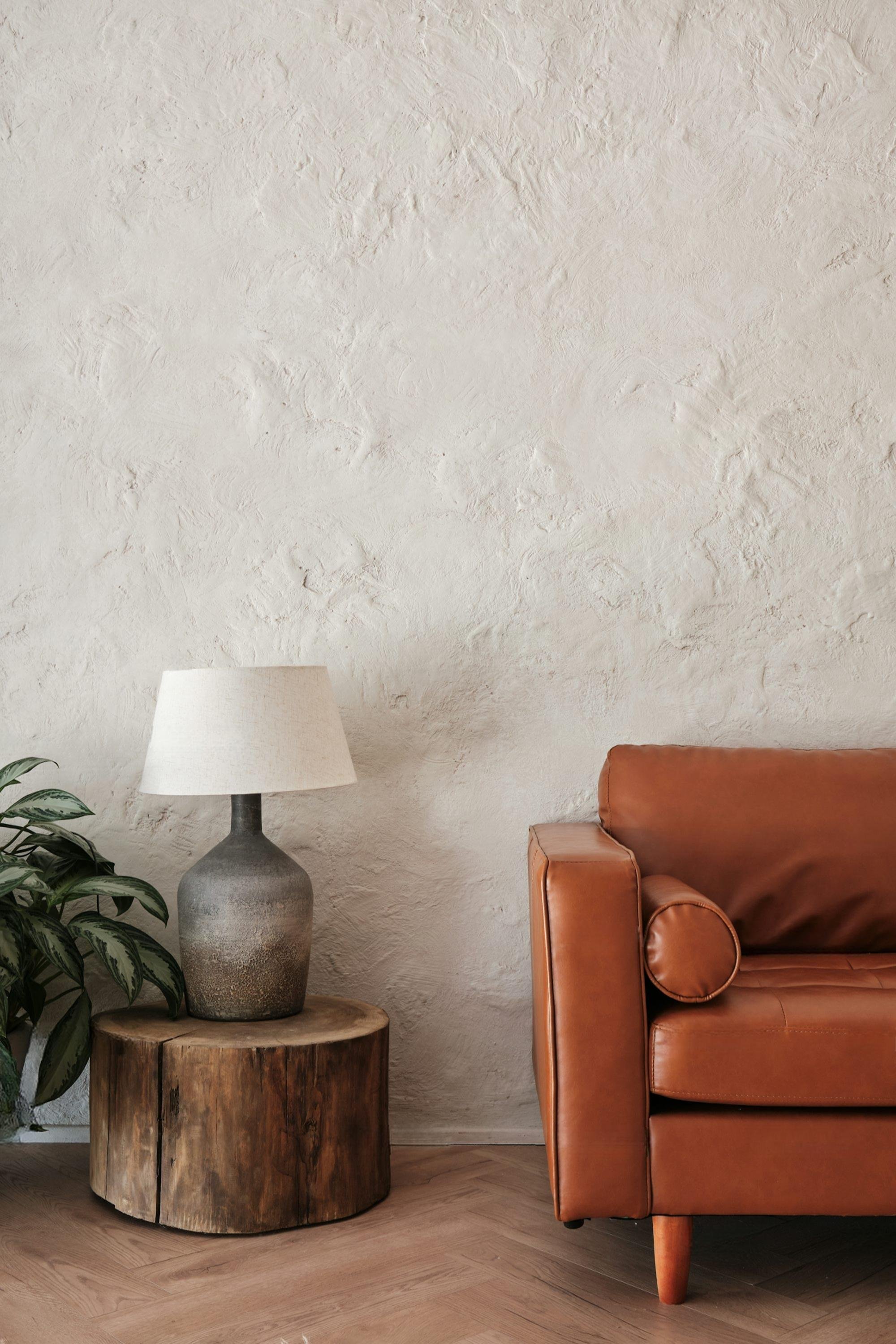 A living room corner with a leather sofa, a wooden side table, a ceramic table lamp with a white shade, and a green leafy plant on the left, against a textured beige wall and wooden flooring.