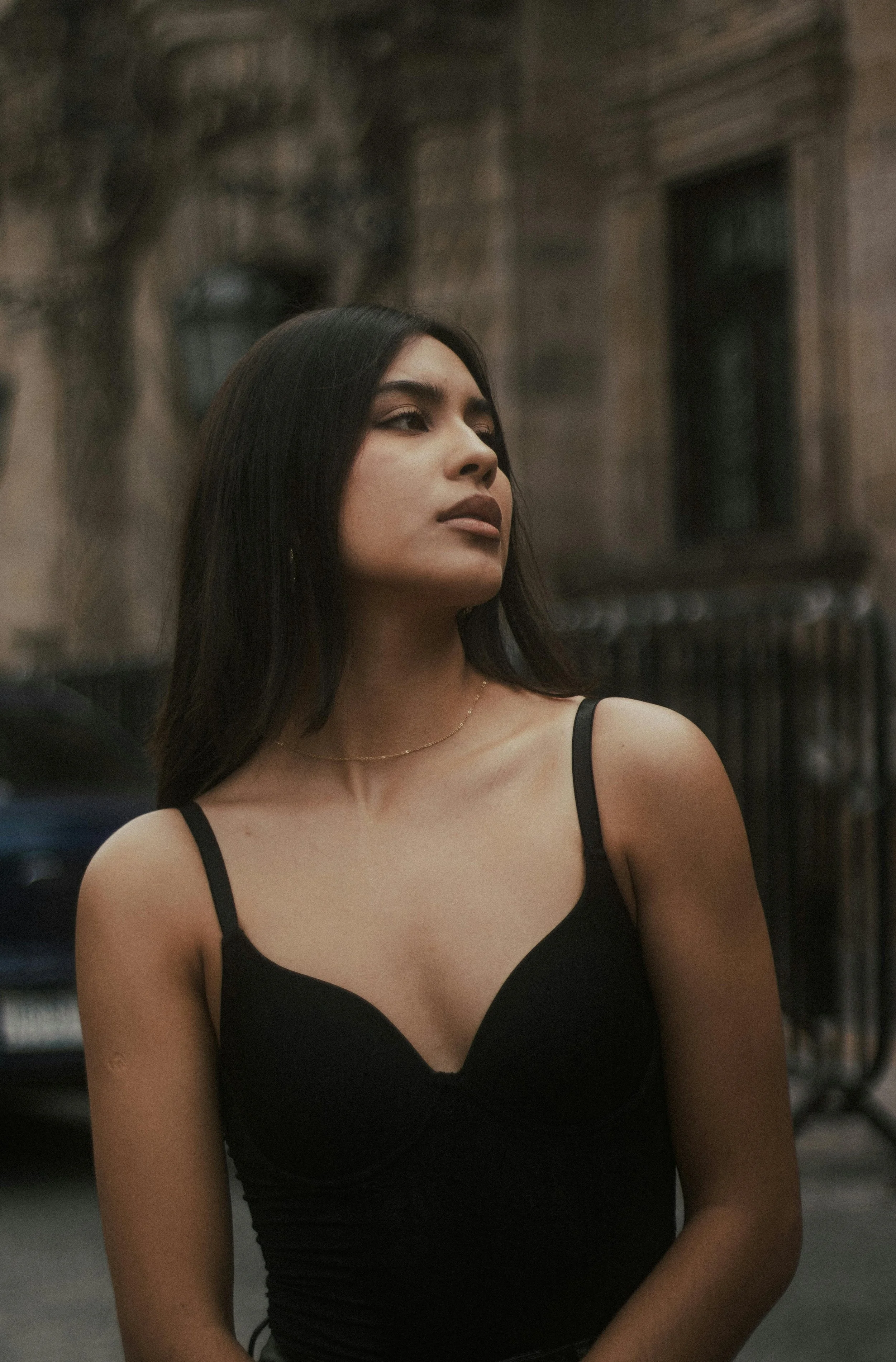 A young woman with long dark hair wearing a black spaghetti strap top, standing outdoors in front of a building with a metal fence and bare tree branches.