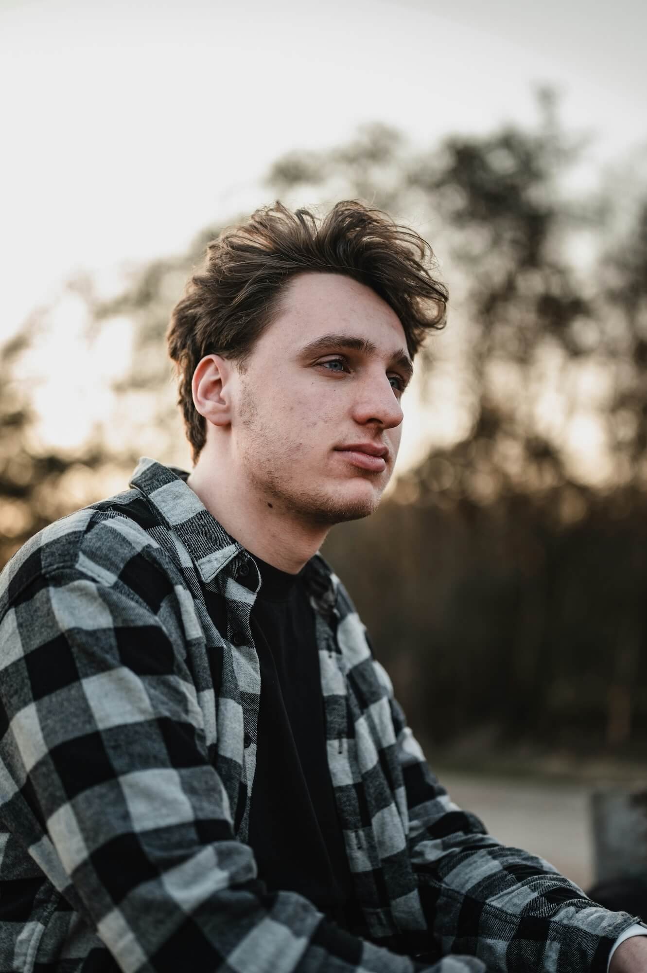 A young man in a black and white checkered shirt sitting outdoors during sunset, looking to the side with a calm expression, with blurred trees in the background.