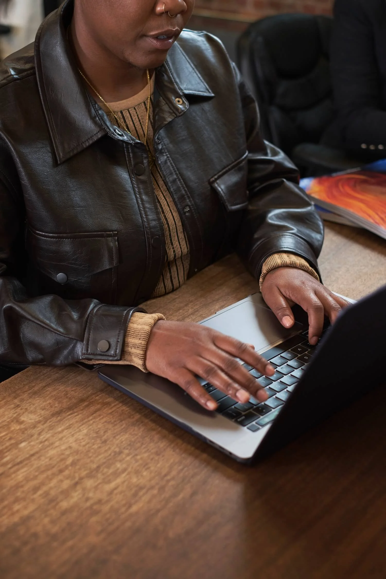 A person wearing a leather jacket and a striped shirt typing on a laptop at a desk.