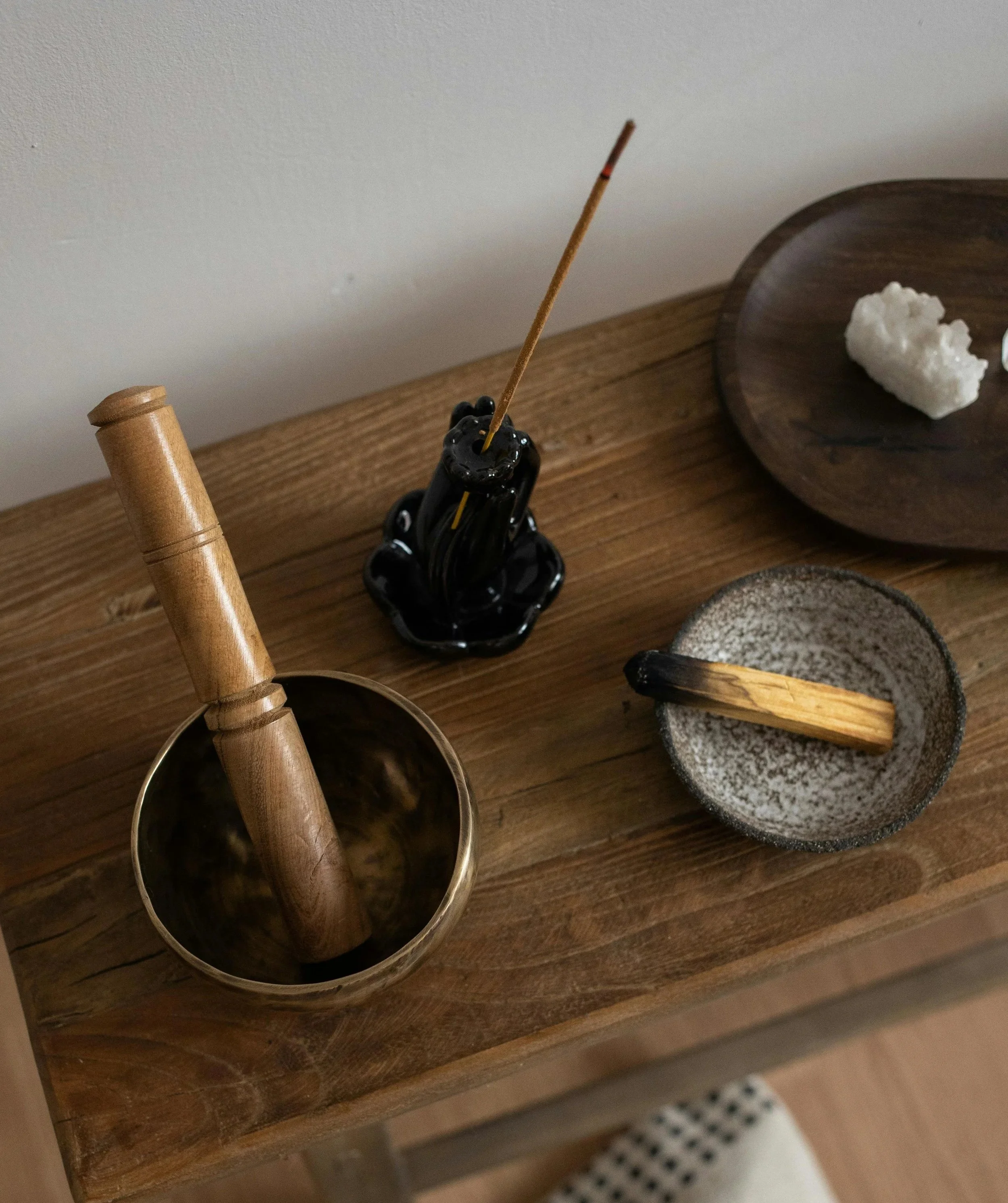A wooden table with black, gray, and wooden objects including a black incense holder with incense, a pestle in a metallic mortar, a wooden stick in a small gray bowl, and a wooden tray with a white object.