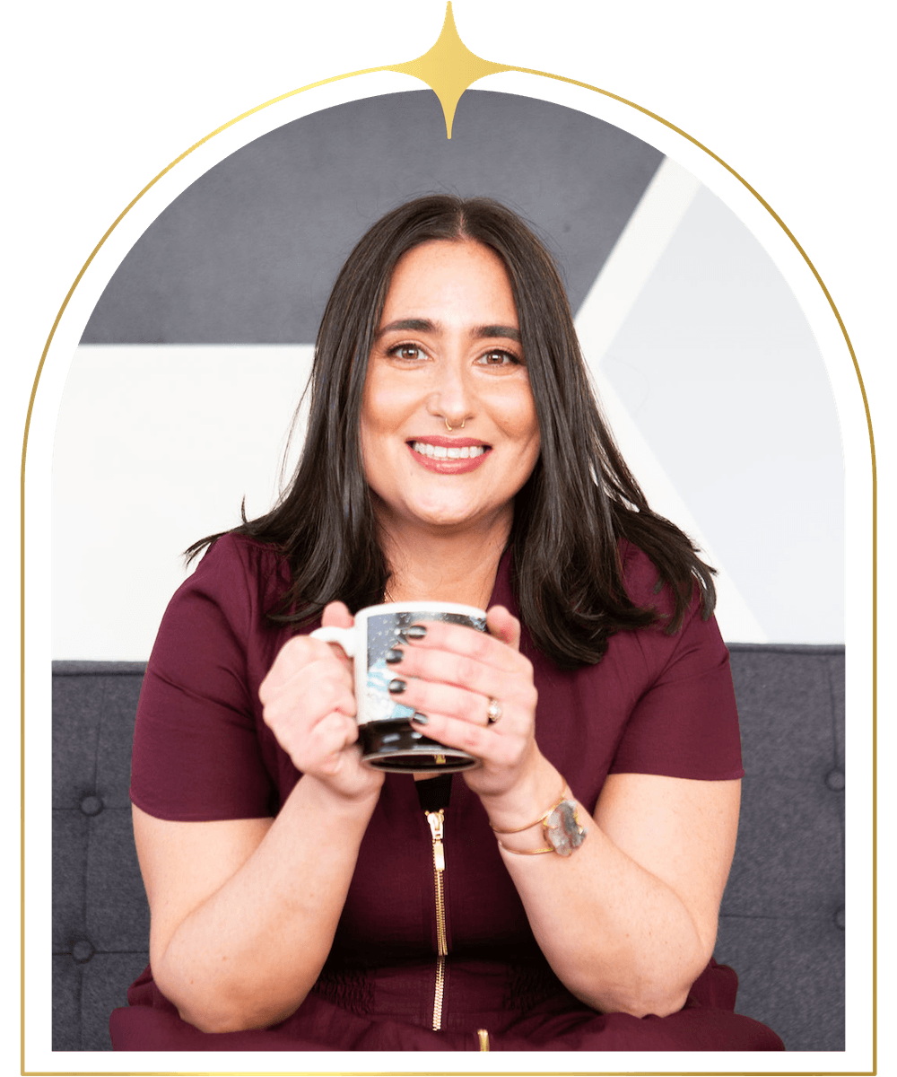 A woman with dark brown hair, smiling, holding a mug, sitting on a gray couch, with an abstract black, white, and gray background.