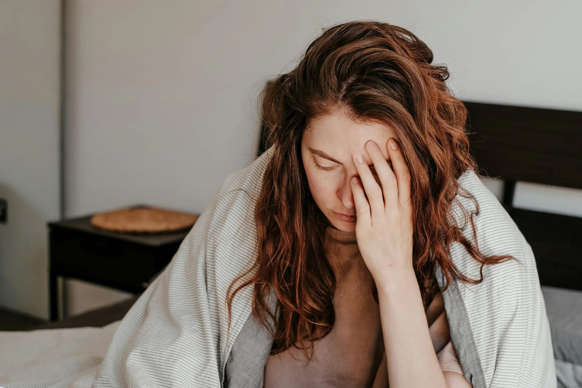 A young woman with wavy red hair sitting on a bed, holding her forehead with her hand, appearing stressed or worried.