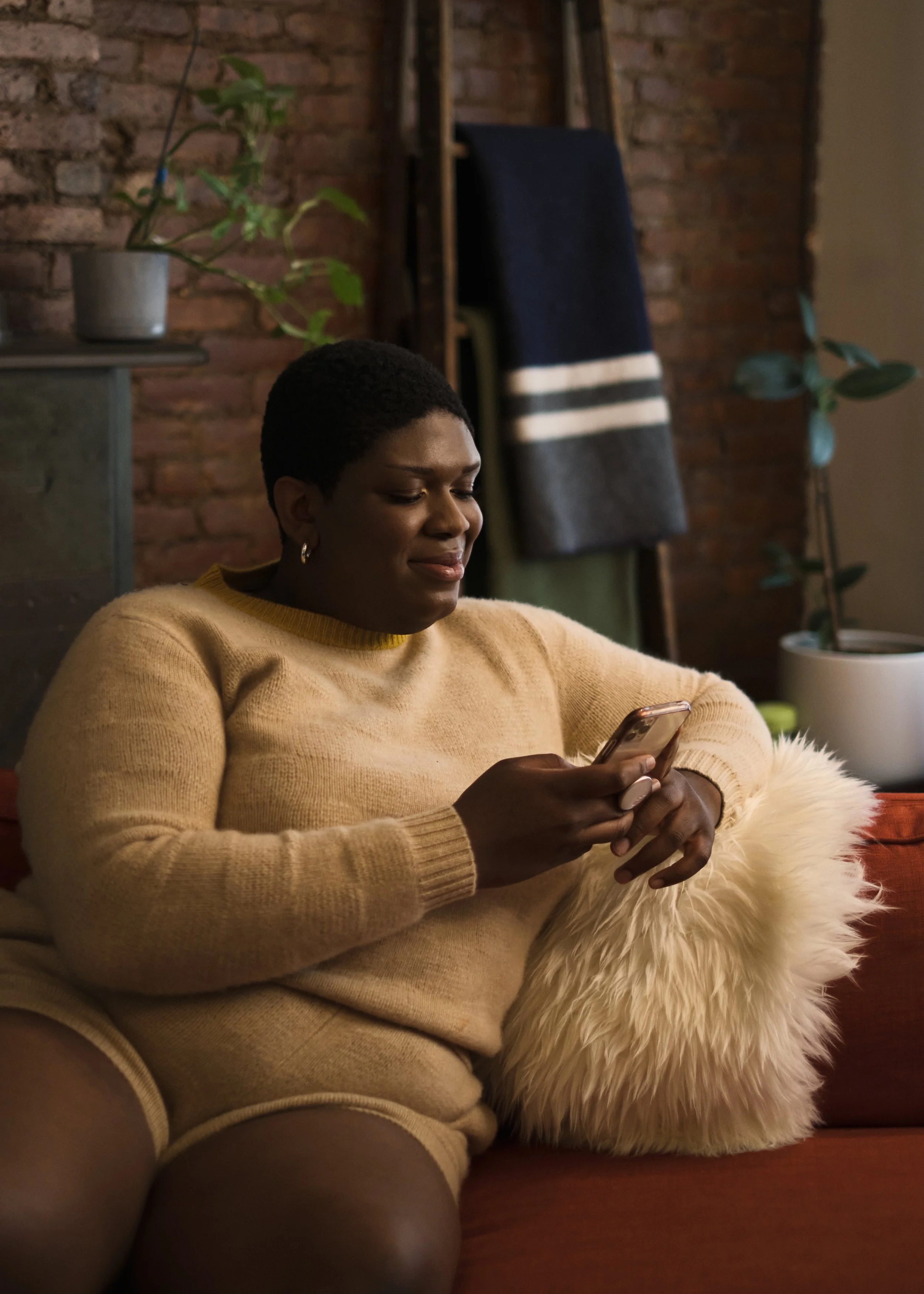 A woman sitting on a sofa with a fluffy pillow, looking at her phone with a smile, in a cozy room with a brick wall and potted plants.