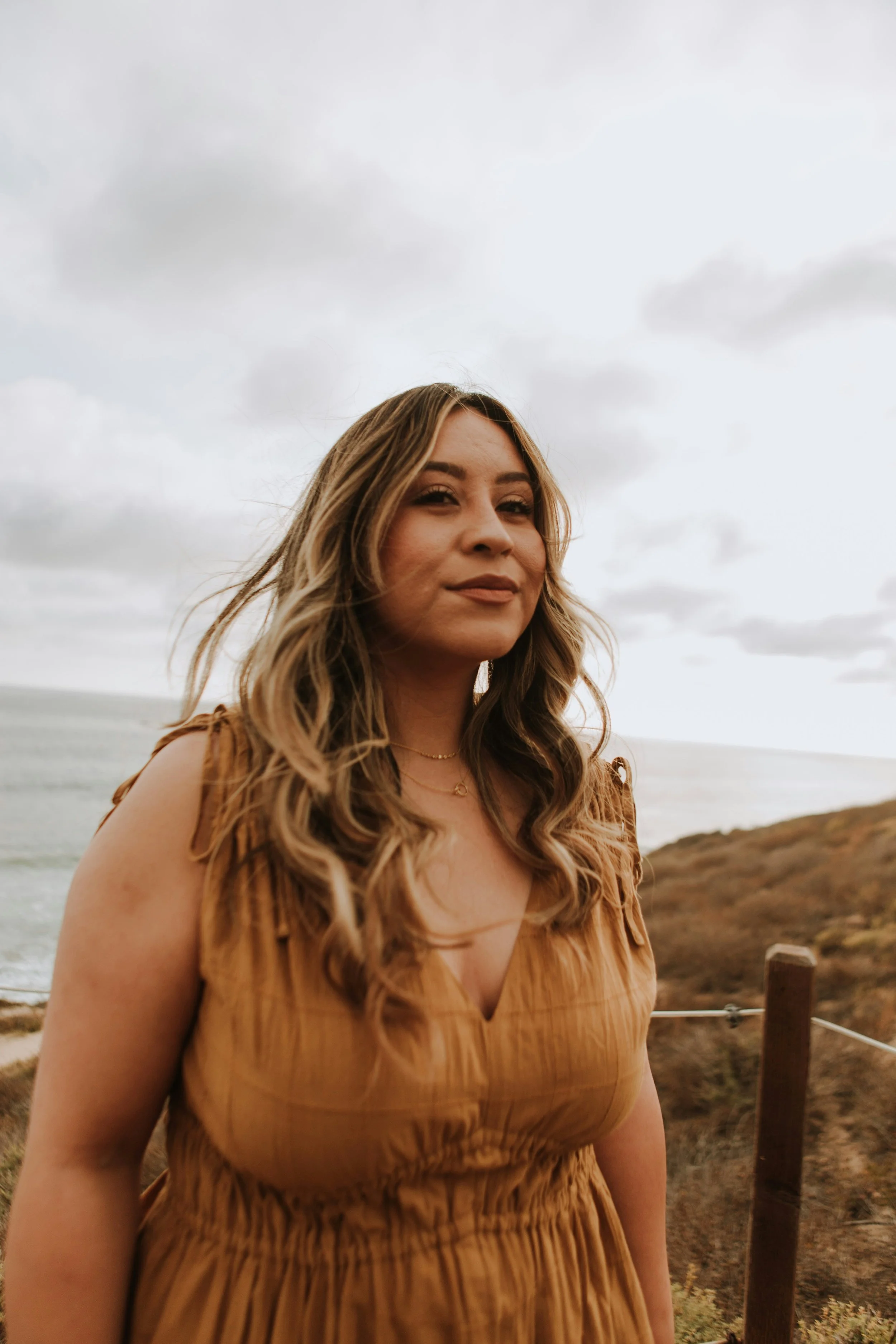 A woman with wavy, shoulder-length hair standing outdoors on a cloudy day, wearing a sleeveless, tan-colored dress, with a beach and hillside in the background.