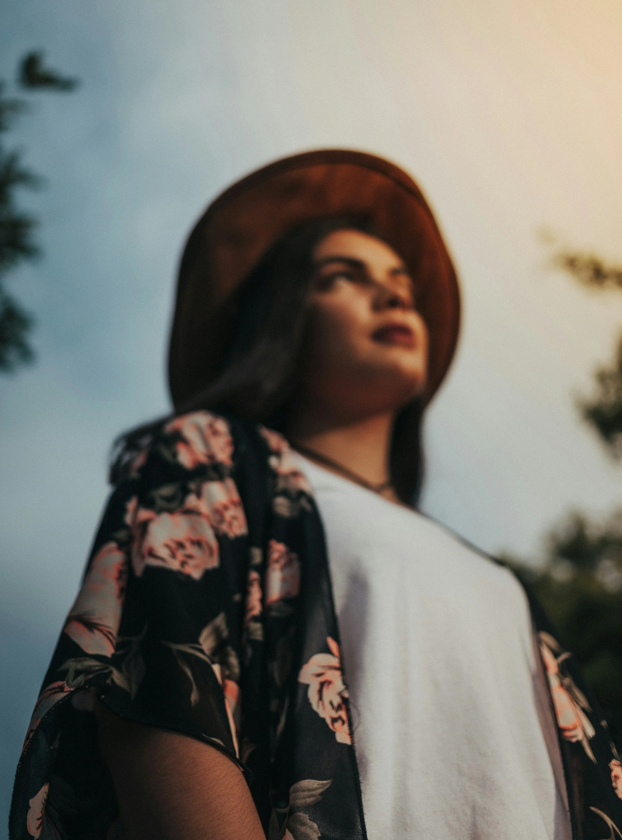 A woman wearing a floral kimono, white t-shirt, and wide-brimmed hat looking upwards outdoors during sunset.