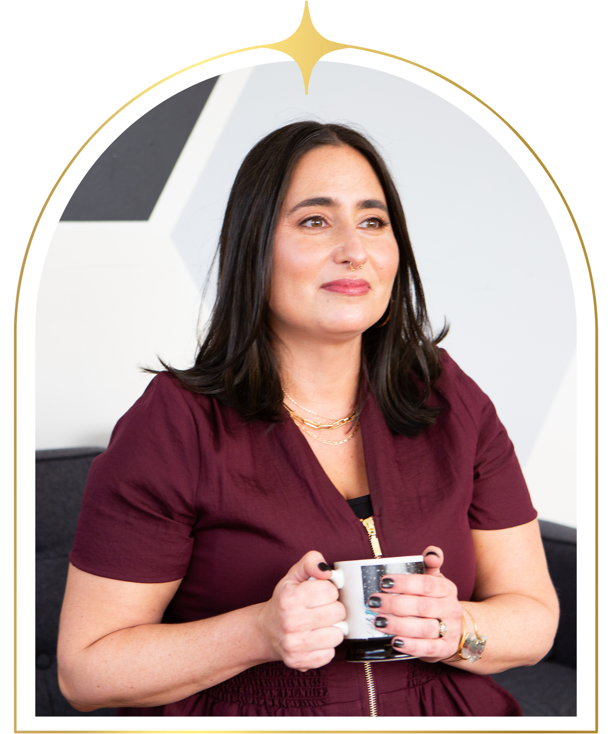 A woman with dark hair is sitting and holding a mug, wearing a burgundy top and layered necklaces, with a neutral expression and a geometric patterned background.