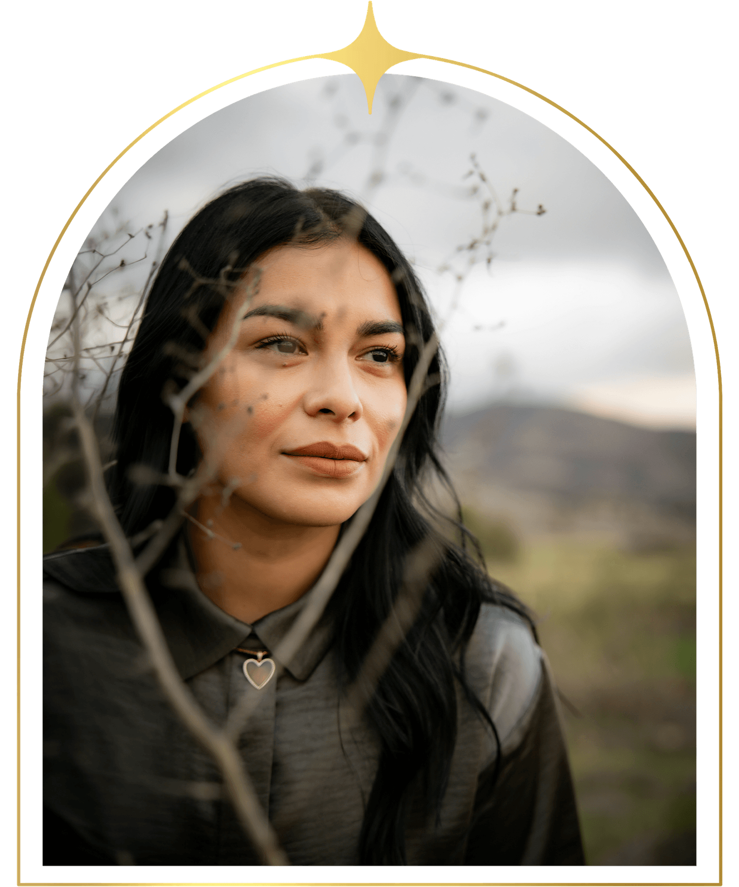 A woman looking through a natural frame created by tree branches, outdoors with mountains in the background.
