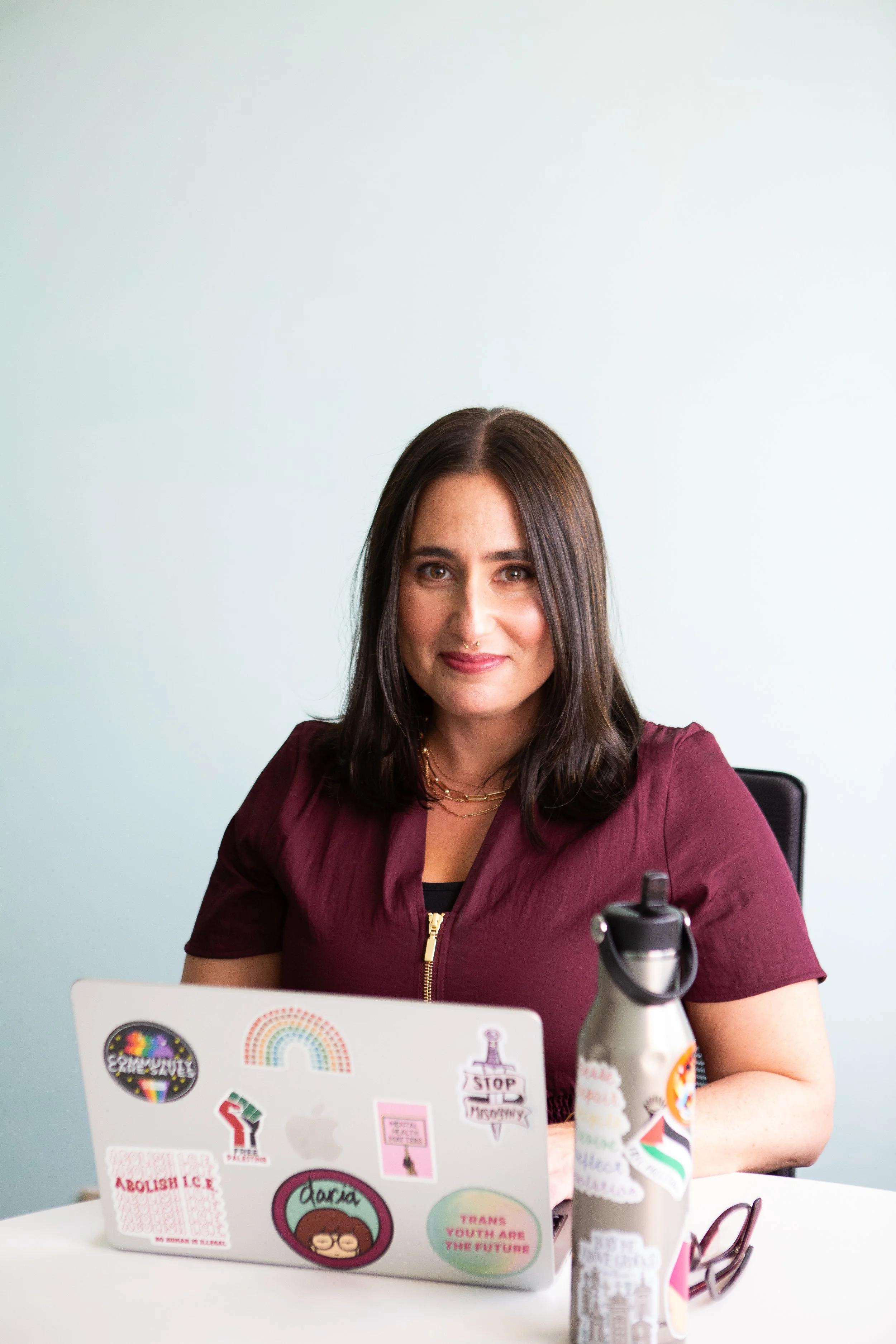 A woman with dark hair sitting at a desk, smiling, with a laptop covered in LGBTQ+ stickers, a water bottle, and glasses on the desk, against a light-colored background.