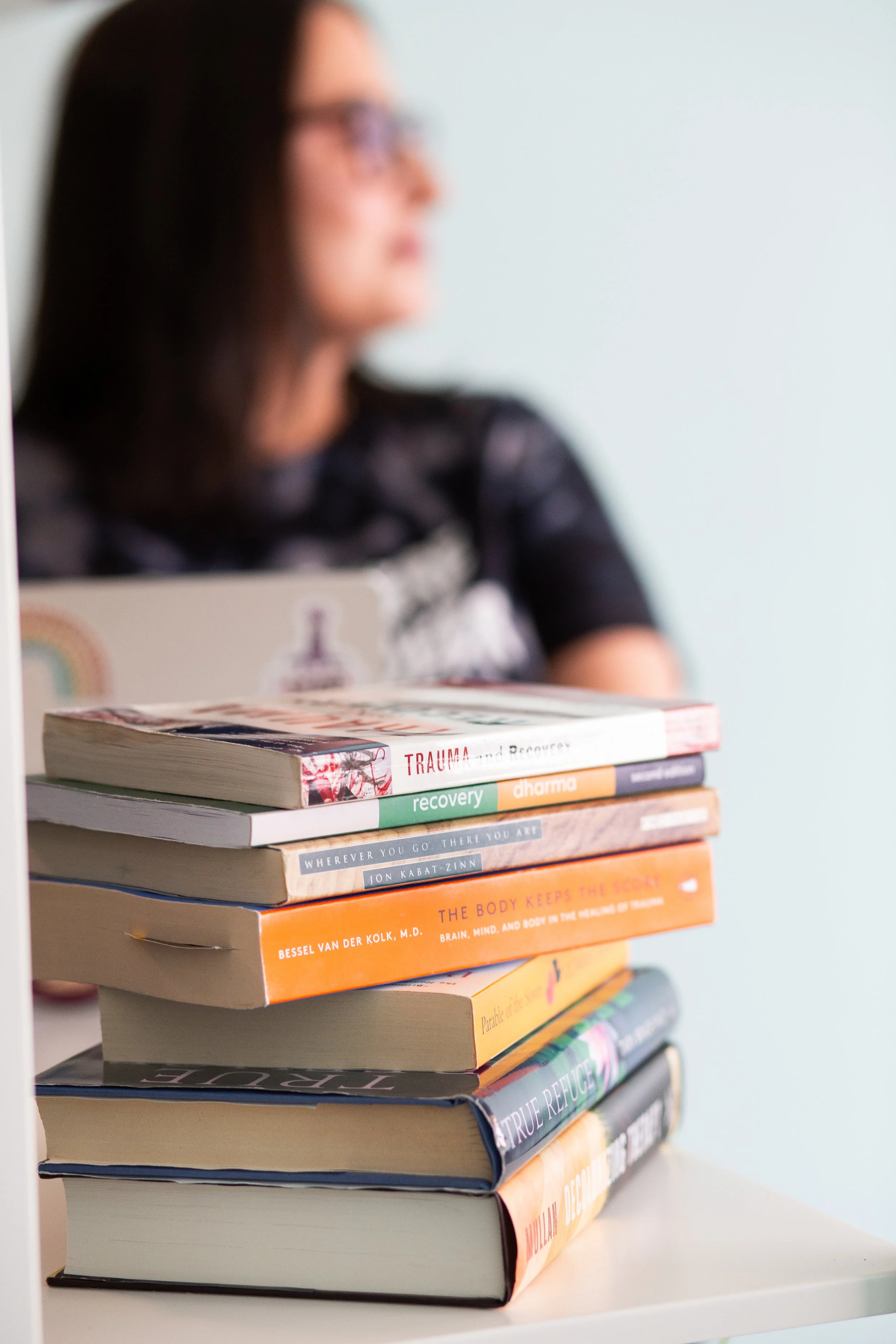 A stack of books on a table with a woman in the background, slightly out of focus, wearing glasses and a black patterned shirt.