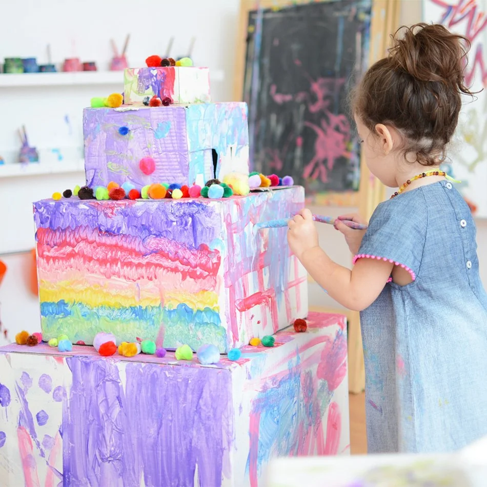 A young girl with brown curly hair, wearing a blue dress with pink pom-pom trim, is painting a colorful, multi-tiered cake sculpture with a brush in a classroom or art studio. Kids art classes, open ended creative workshops located in Kingston, Tasma