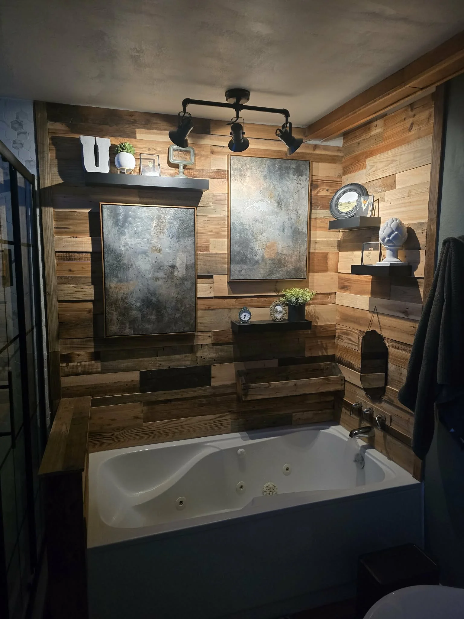 Bathroom with a white jacuzzi tub, wooden wall paneling, shelves with decorative items, clock, plant, and ceiling light fixtures.