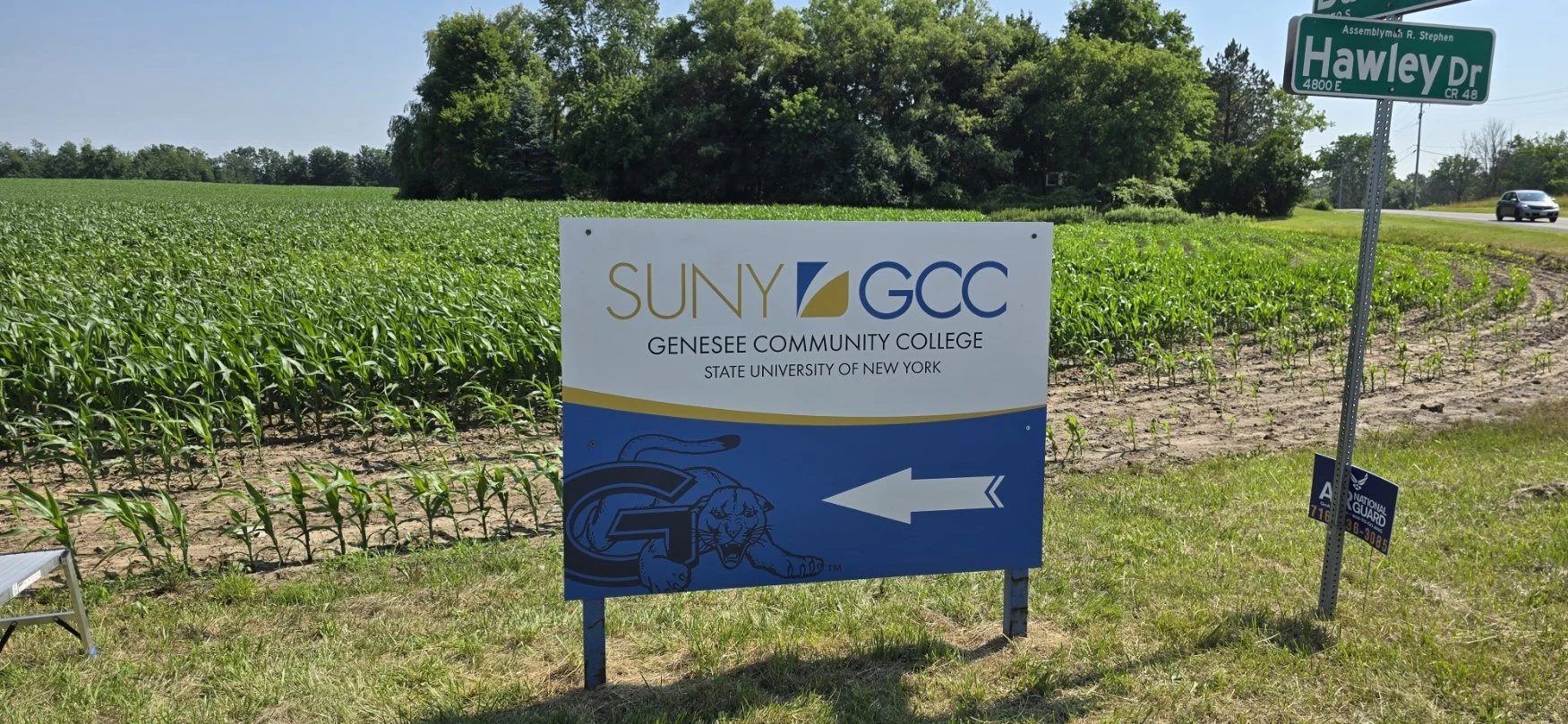 A sign indicating the direction to Genesee Community College, part of SUNY-GCC, located on Hawley Drive, with a cornfield and trees in the background.