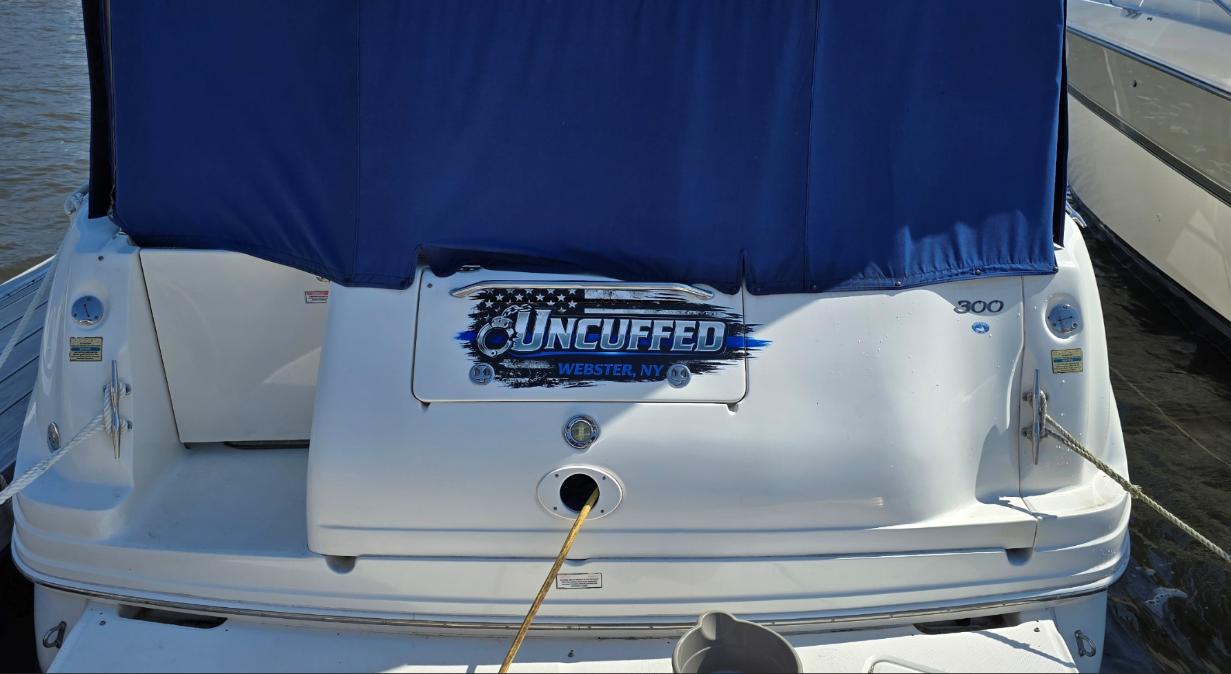 Back of a white boat with a blue cover, displaying a decal that says 'Uncuffed, Webster, NY' , tied to a dock.