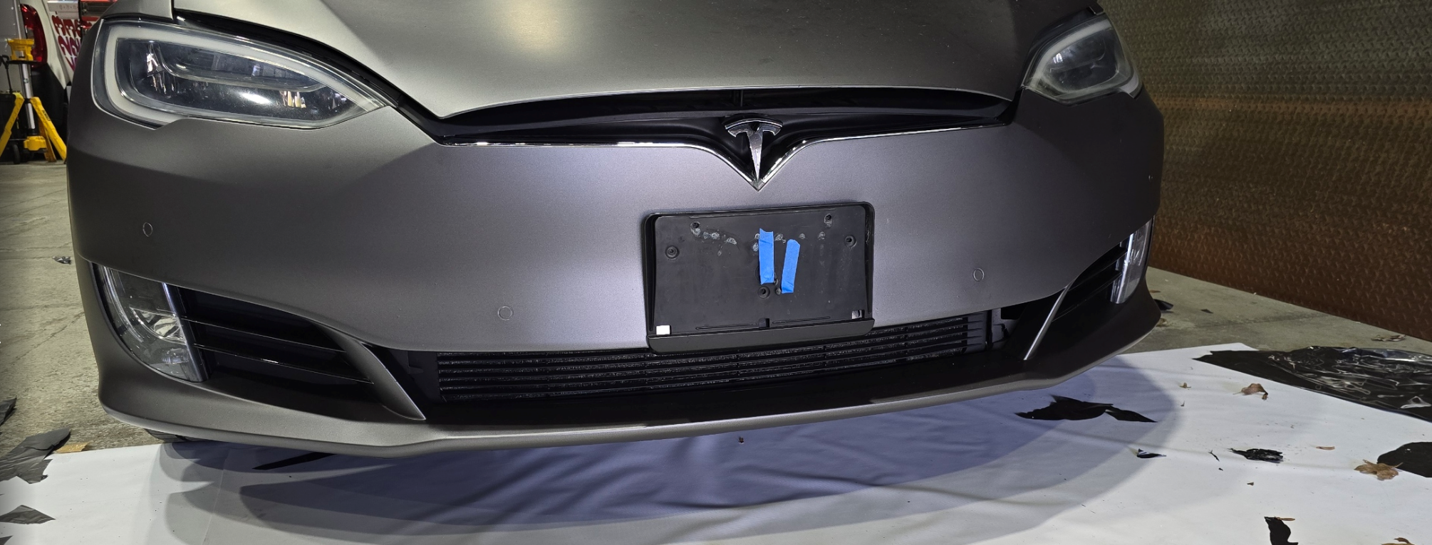 Front view of a silver Tesla Model S with a black license plate holder and no license plate, parked indoors on a white surface with black and brown debris, and a brown wall with textured pattern in the background.