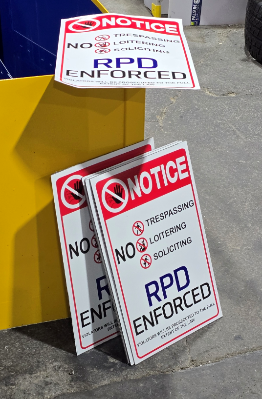 Stack of security notices and a full-sized notice sign on a concrete floor, with yellow metal barricade in the background. The signs indicate no trespassing, loitering, or soliciting, and mention RPD enforced.
