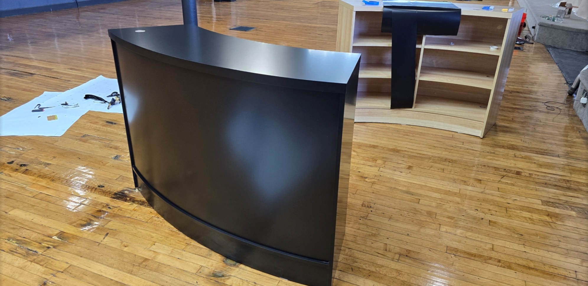 Disassembled black and wood-colored curved curved office desk and a wooden bookshelf in a room with a wooden floor, tools, and packaging materials.