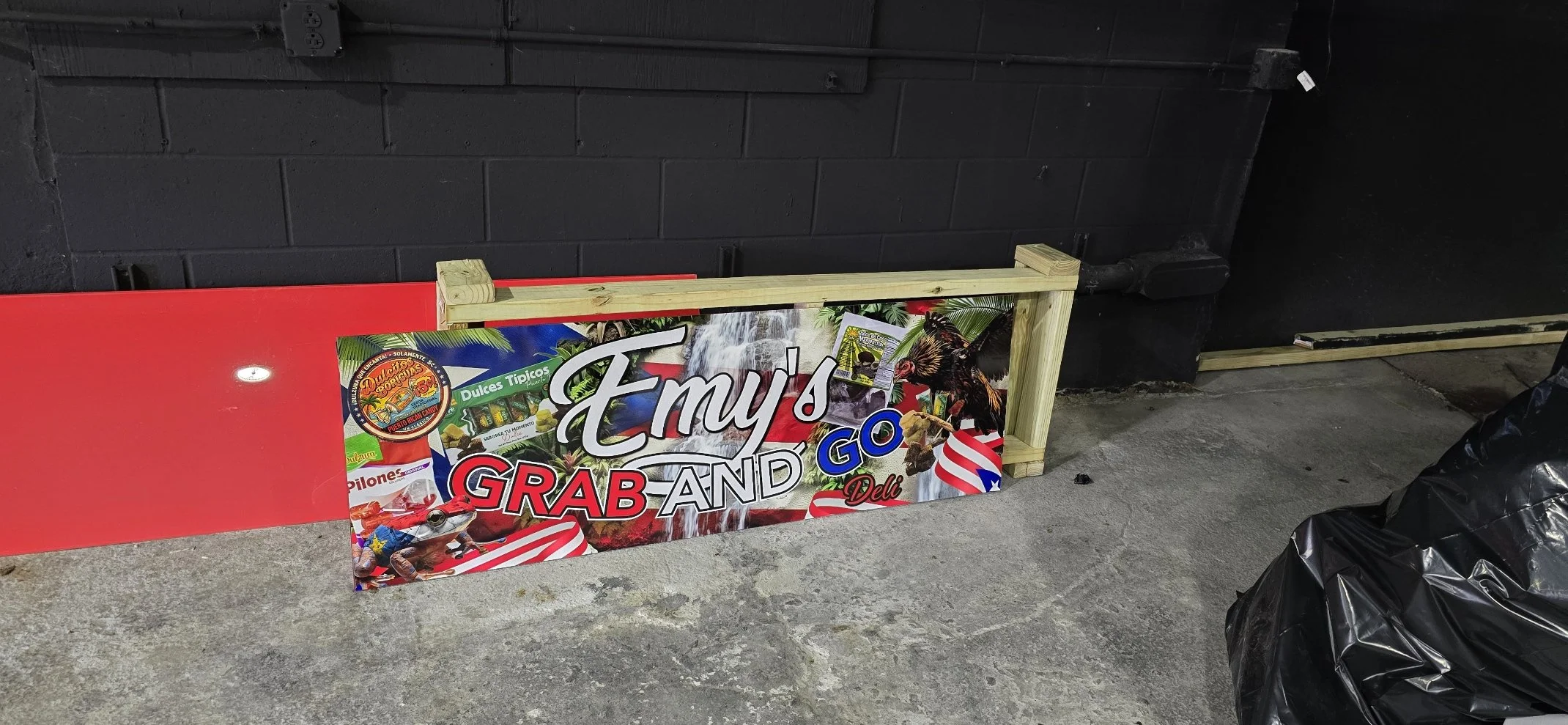 A colorful sign that reads 'Emy's Grab and Go Deli' with images of tropical fruit, a rooster, and patriotic American flag elements, leaning against a black painted brick wall with a wooden frame partially surrounding it.