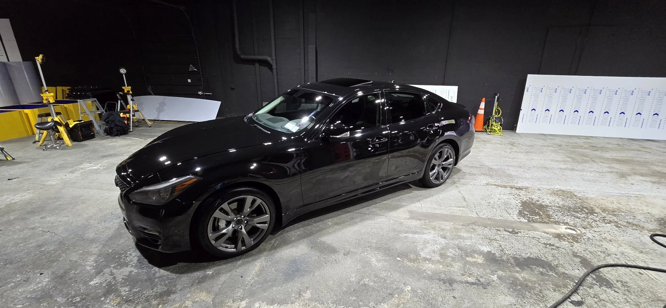 A black sedan car parked inside a dimly lit garage with construction materials and tools on the side, including yellow equipment, white insulation board, and an orange traffic cone.
