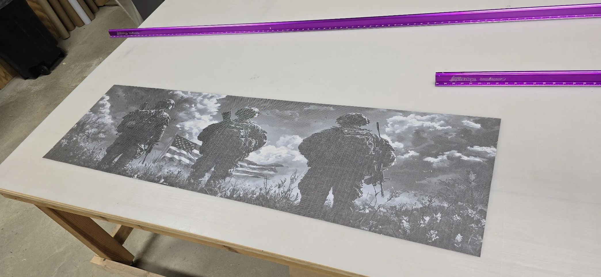 Black and white photo of three soldiers standing outdoors with cloudy sky and trees in background, one holding a flag, on a table covered with mesh-like material, with purple rulers nearby.
