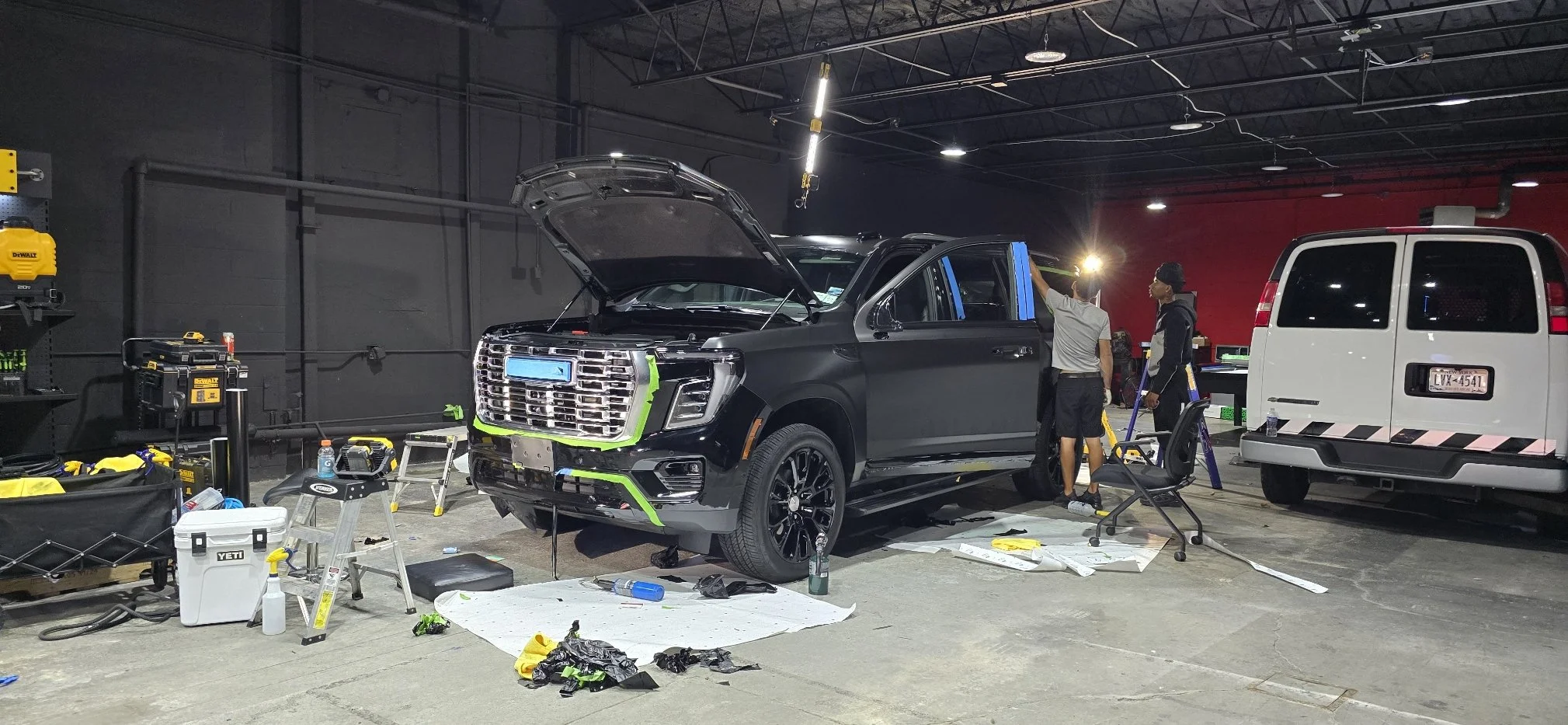 Automotive workshop with a black SUV, possibly a new model, undergoing repairs or modifications. The hood is open, and two men are working near the vehicle. The workspace is equipped with tools and equipment, including a yellow power tool, a cooler, and a workbench. A white van is parked nearby.