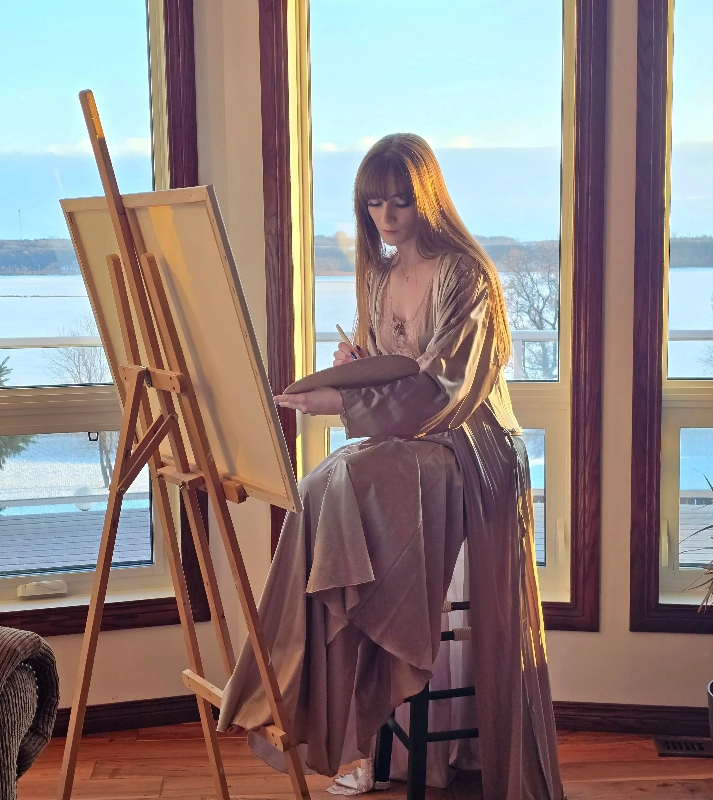 A woman with long reddish brown hair and bangs, wearing a satin dress, sitting by large windows with a view of a river and valley or lake and trees outside. She is painting on a large canvas and easel in her home art studio in Manitoba
