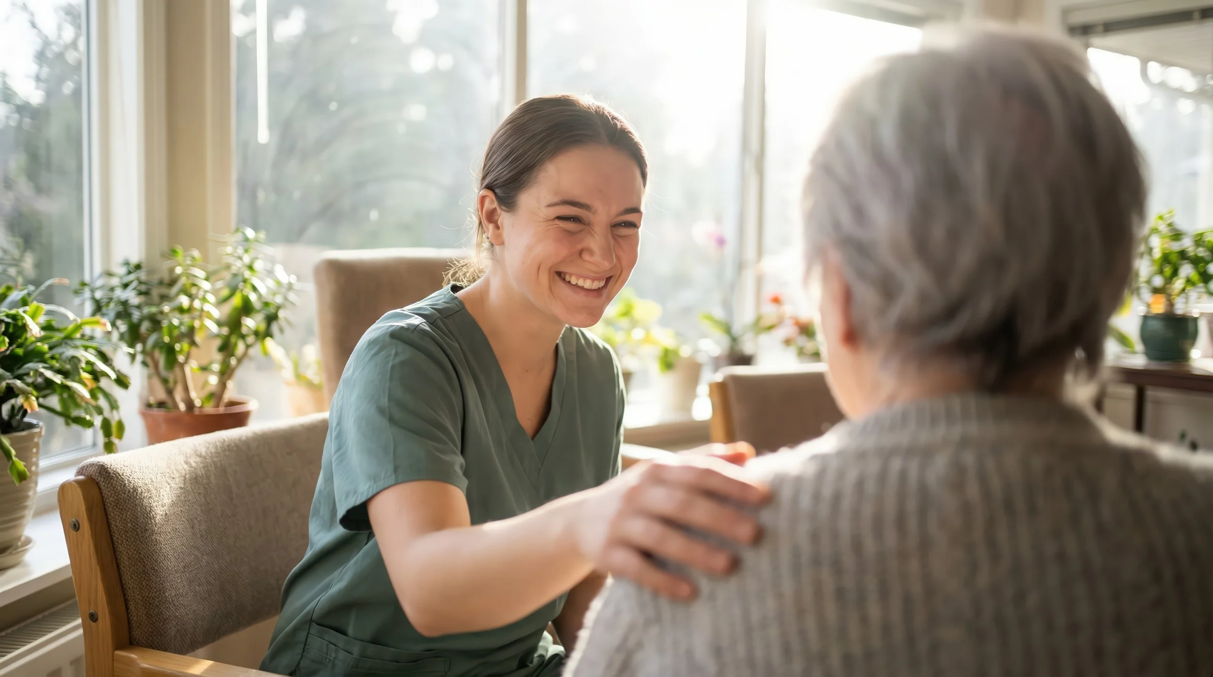 A young woman in medical scrubs smiling and holding hands with an elderly man with gray hair, sitting at a table with potted plants behind them in a sunlit room.