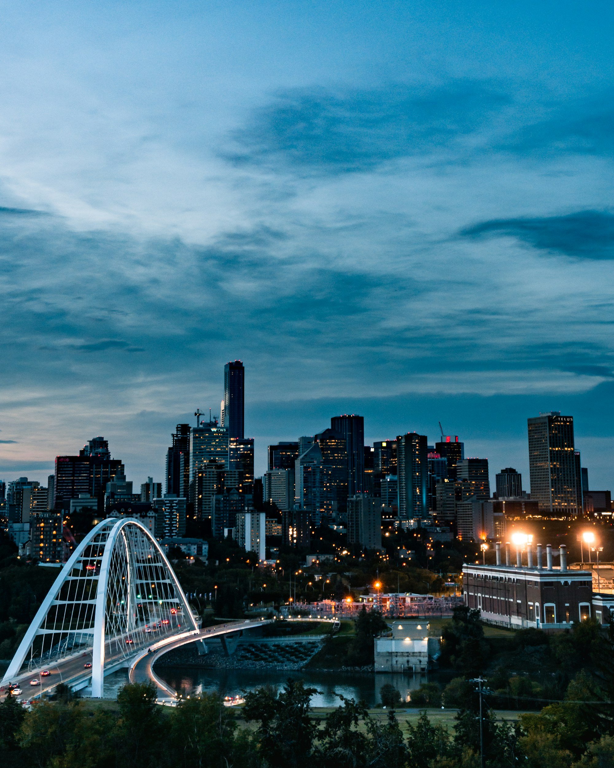 The image shows a city skyline at dusk with tall skyscrapers and buildings. A distinctive white arched bridge crosses a body of water in the foreground, with cars traveling on it. The sky is partly cloudy with blue and gray tones, and city lights are starting to turn on.