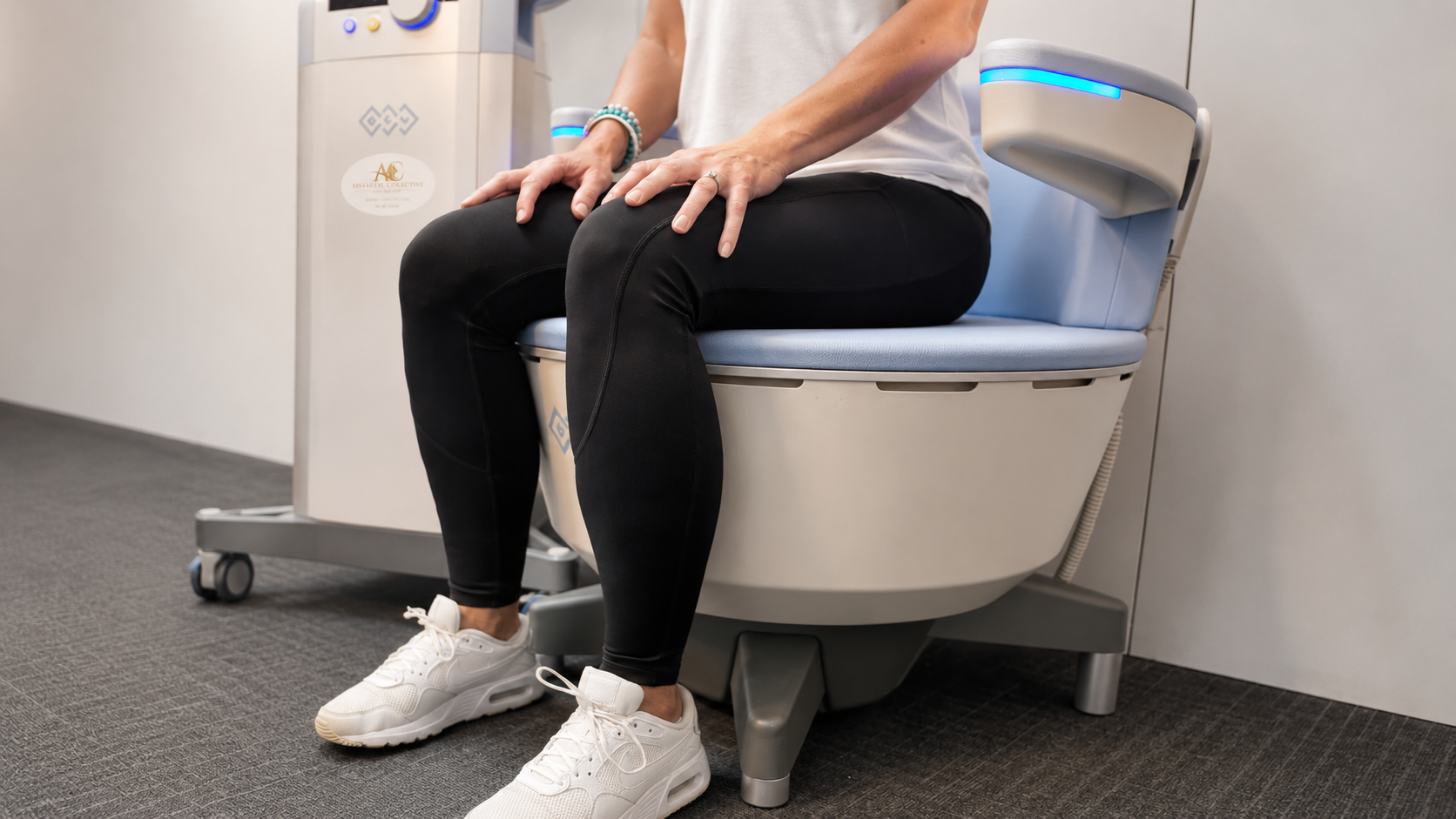 A person sitting on a modern toilet with a remote control panel on the side. The person is wearing black leggings, white sneakers, and has bracelets on their wrist.