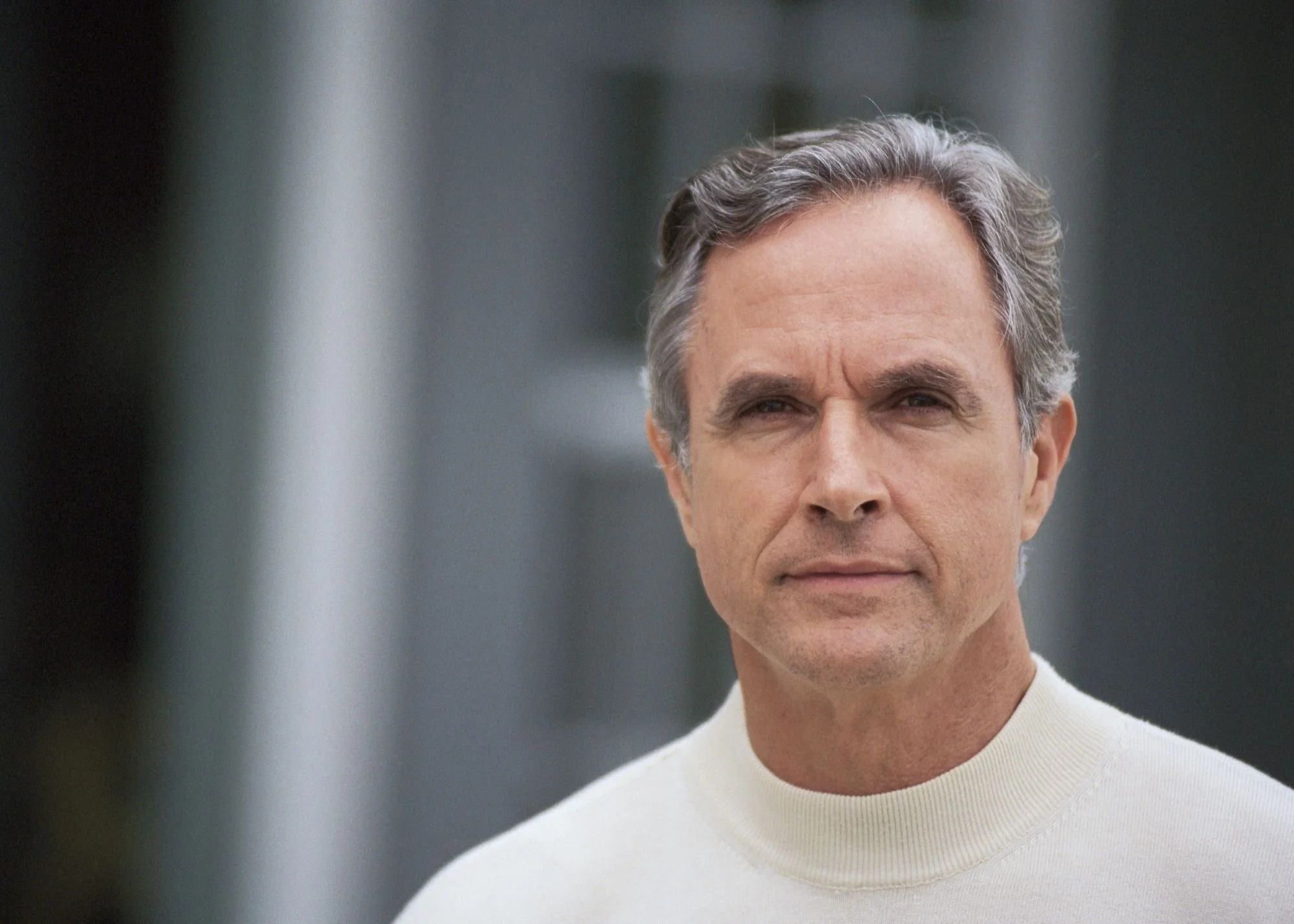 Close-up of a middle-aged man with gray hair wearing a white sweater, looking directly at the camera.