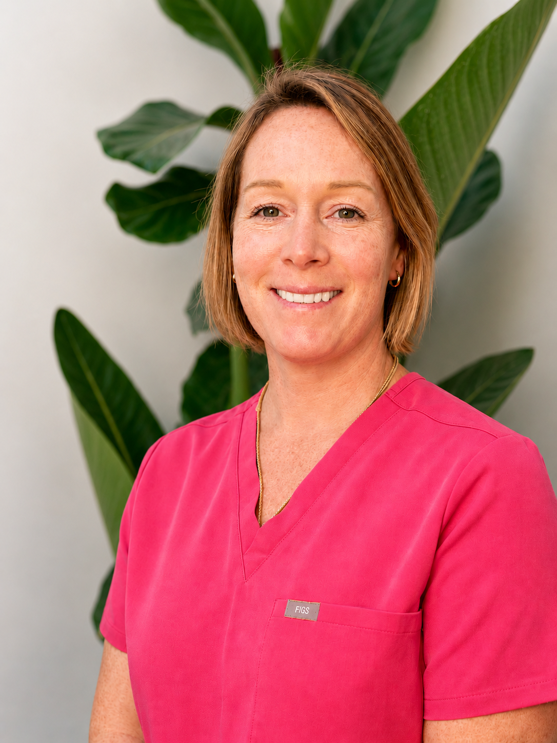 Aesthetics Collective owner Jarnah Snee, wearing a pink medical scrub top, standing in front of large green leaves.