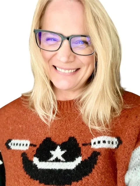 A woman with blonde hair and glasses smiling, wearing a western sweater with a black cowboy hat and a white star.