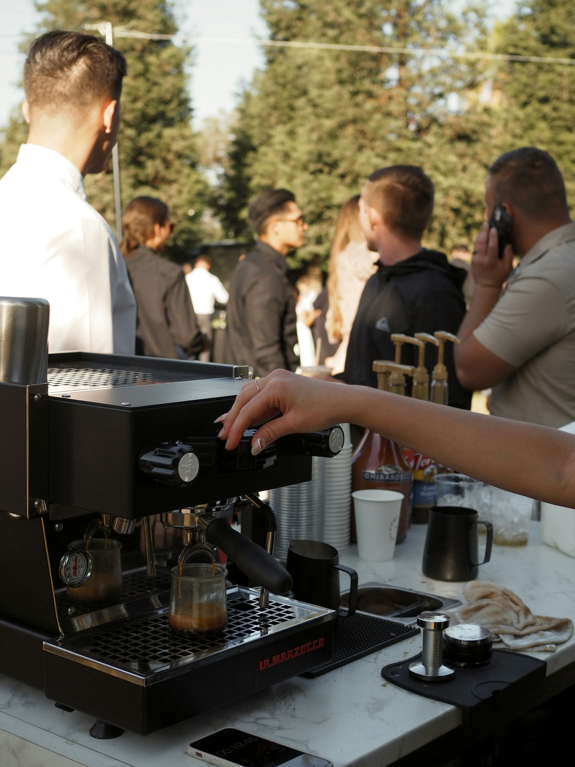 Close-up of a barista's hand adjusting a coffee machine at an outdoor event, with people in the background.