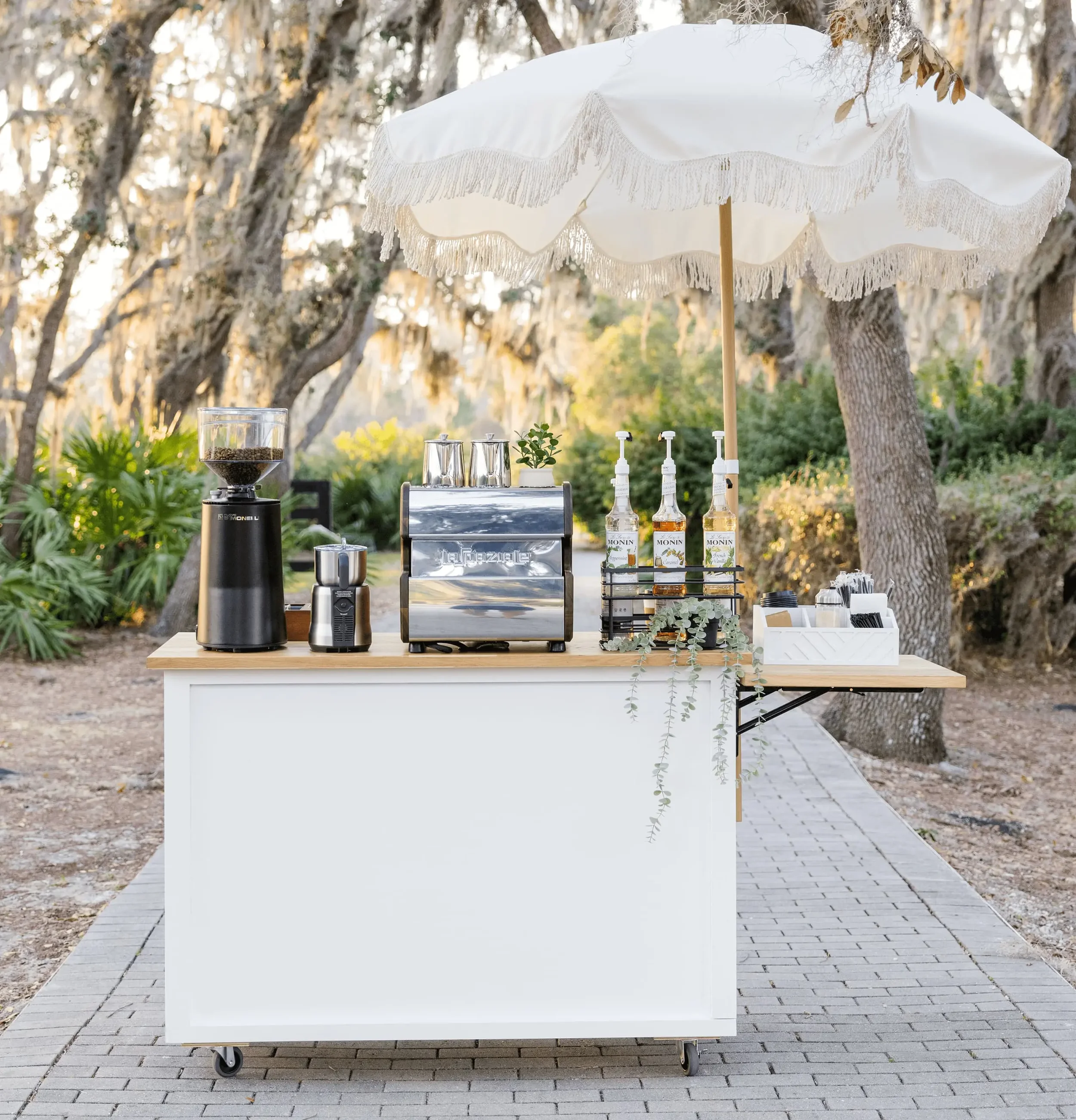 Coffee station setup outdoors with a coffee grinder, espresso machine, and bottles of Monin flavored syrup under a large white umbrella, surrounded by trees and greenery.