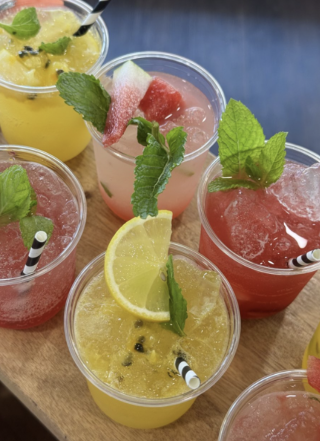 Six colorful fruit drinks in clear plastic cups with lemon and watermelon slices, fresh mint leaves, and striped paper straws on a wooden surface.